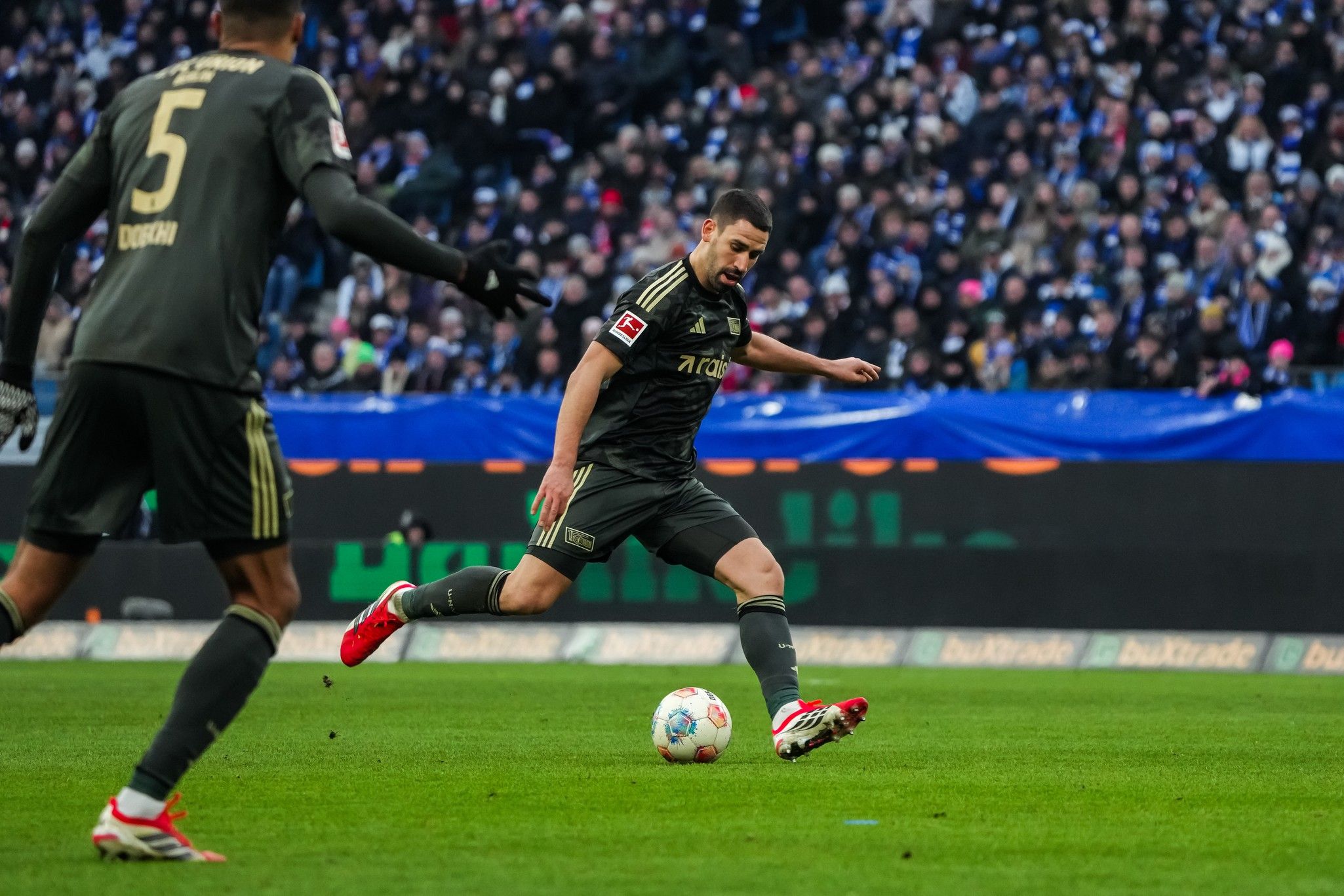 Ein Fußballspieler in grauer Uniform tritt den Ball während eines Spiels vor einem jubelnden Publikum.