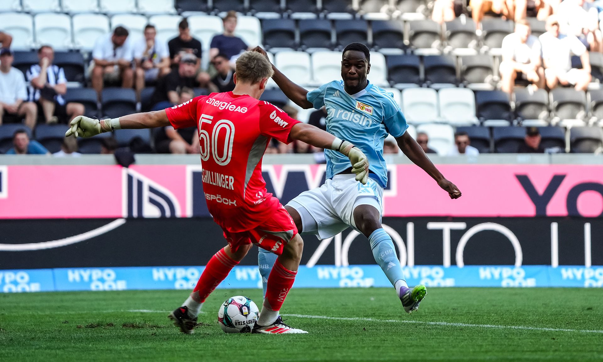 Ein Fußballspieler in blauer Uniform versucht, einem Torwart in roter Uniform auszuweichen, während der Ball in Bewegung ist.