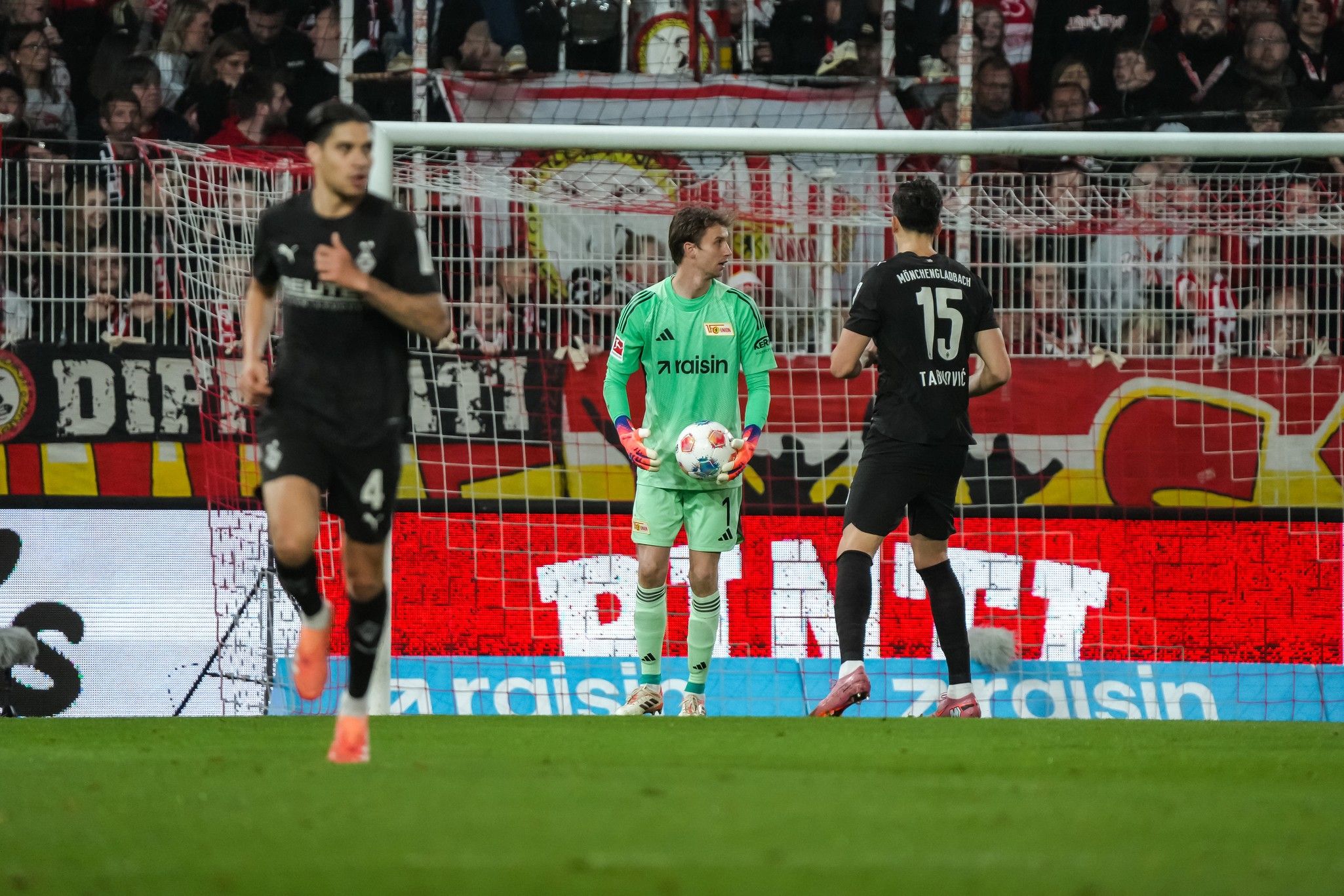 Ein Fußballspiel im Stadion, mit einem Torwart in grüner Ausrüstung und Spielern in schwarzen Trikots im Hintergrund.
