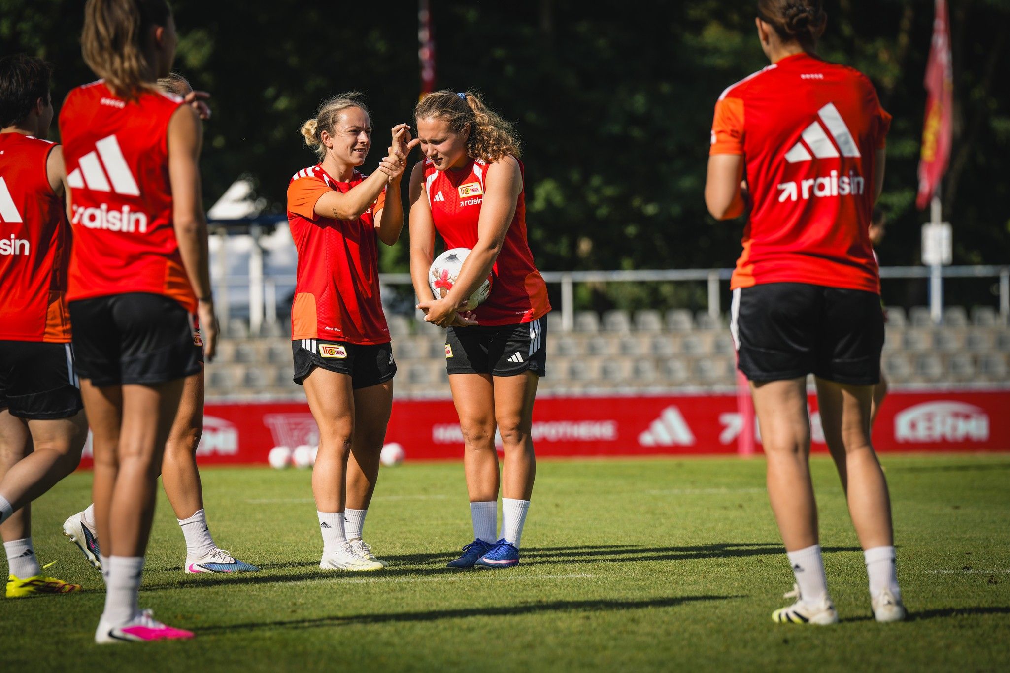 Zwei Spielerinnen in roten Trikots stehen in einem Sportfeld und unterhalten sich, während andere Spielerinnen im Hintergrund trainieren.