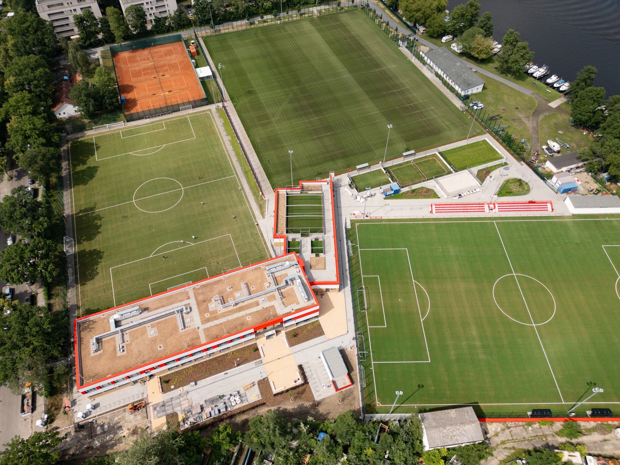 Luftaufnahme eines Sportgeländes mit mehreren Fußballfeldern, einem Tennisplatz und angrenzenden Gebäuden.