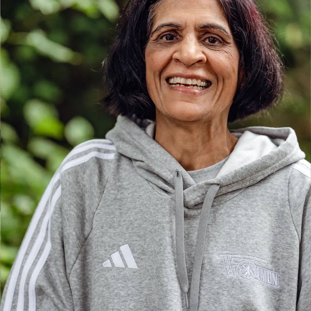 Eine Frau mit einem Lächeln steht vor einem grünen Hintergrund. Sie trägt einen grauen Kapuzenpullover. Der Pullover hat weiße Streifen.
