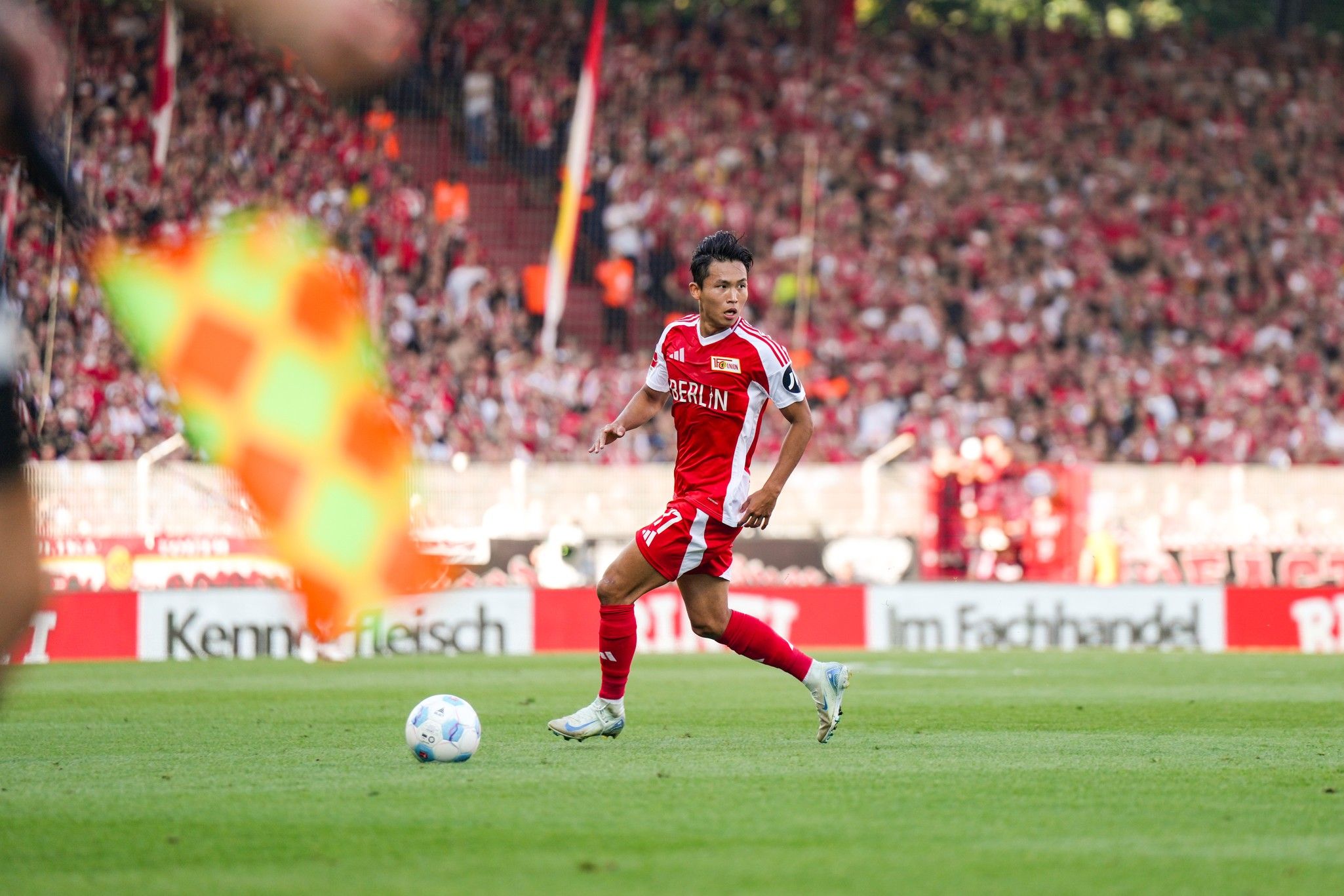 Ein Fußballspieler in rotem Trikot läuft über das Spielfeld, während Zuschauer im Hintergrund jubeln.