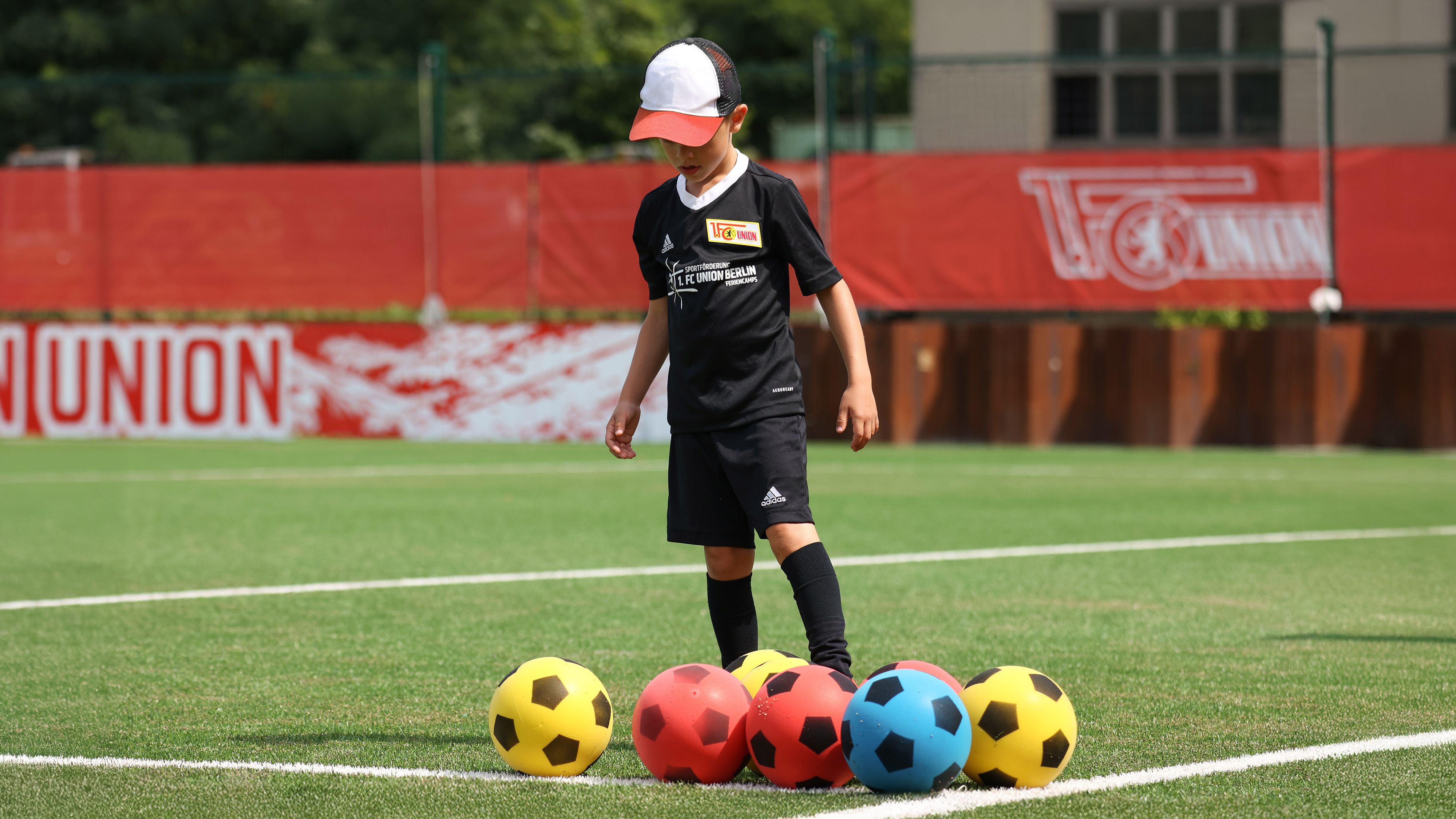 Ein Junge in schwarzer Sportkleidung steht auf einem Fußballfeld und betrachtet mehrere bunte Fußbälle in Rot und Gelb.