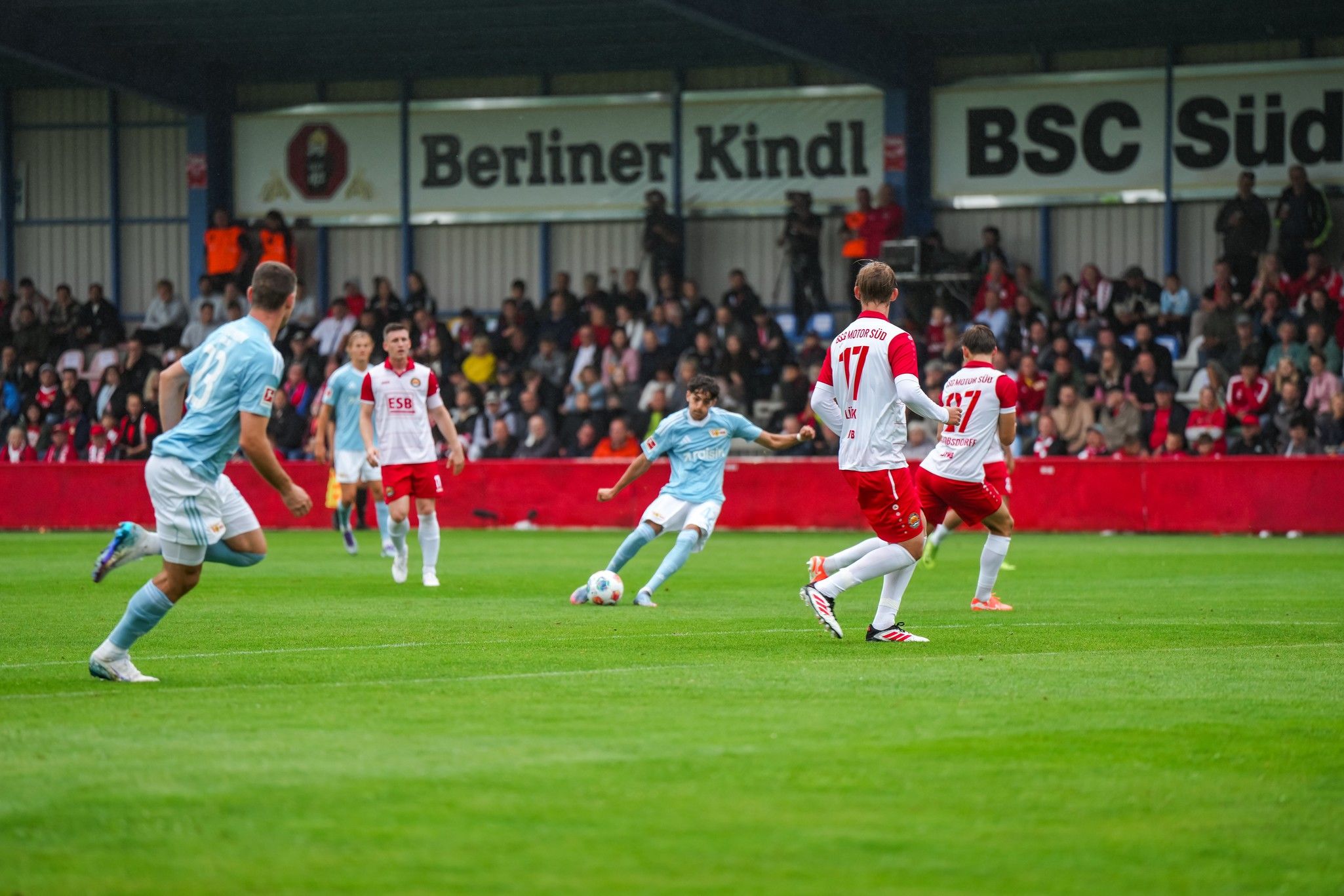 Spielszene im Fußball mit Spielern in blauen und roten Trikots auf einem Rasenplatz und einer zuschauenden Menge im Hintergrund.