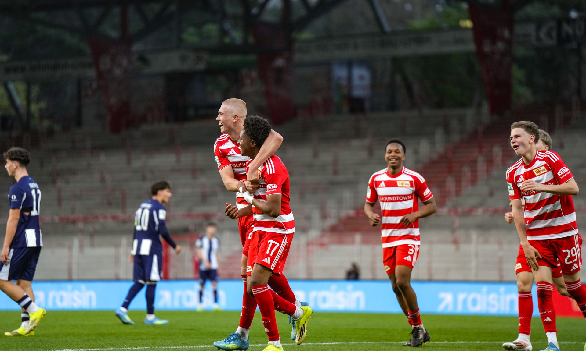 Fußballspieler feiern ein Tor auf dem Spielfeld, während gegnerische Spieler im Hintergrund stehen.