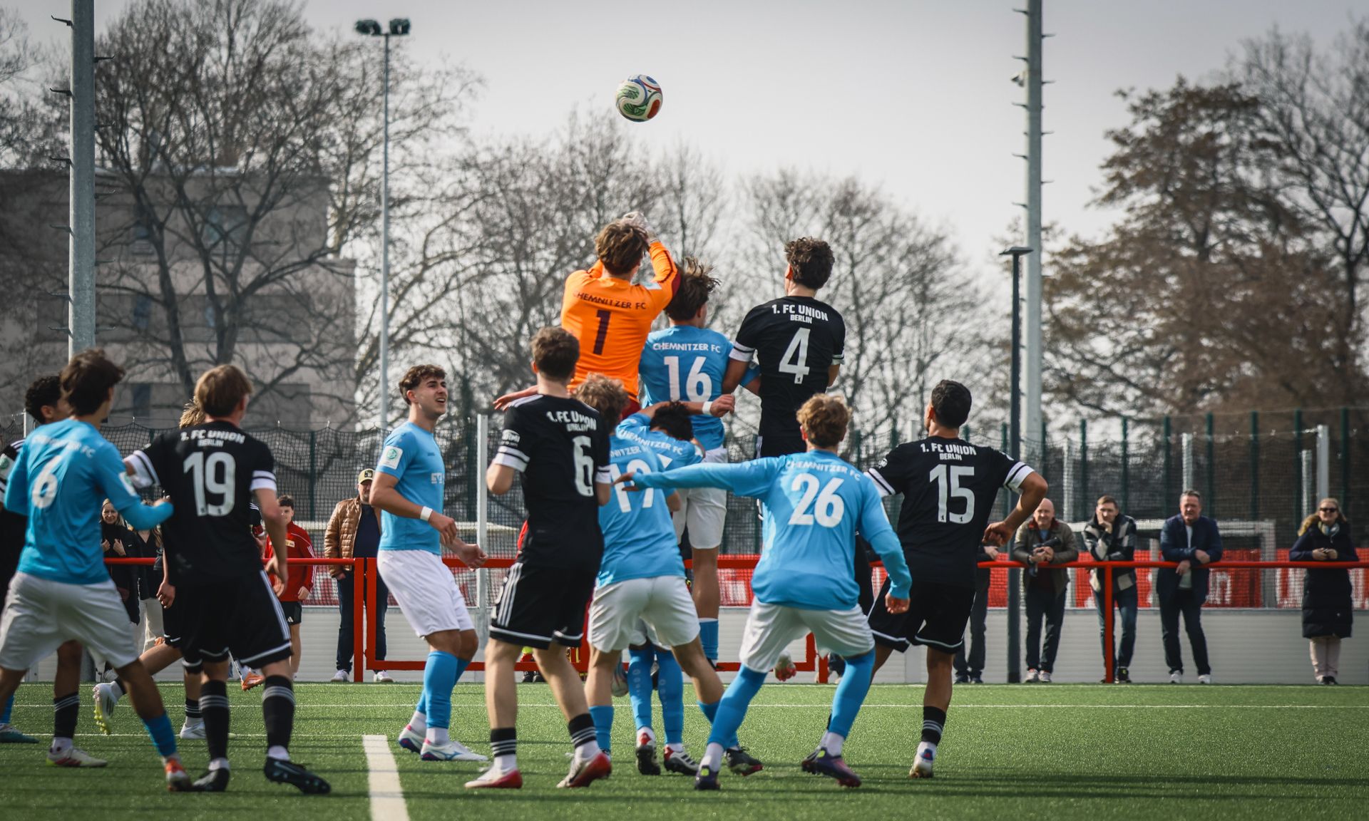 Spielszene eines Fußballspiels, bei dem der Torwart den Ball hoch über Spieler in schwarzen und blauen Trikots springt.