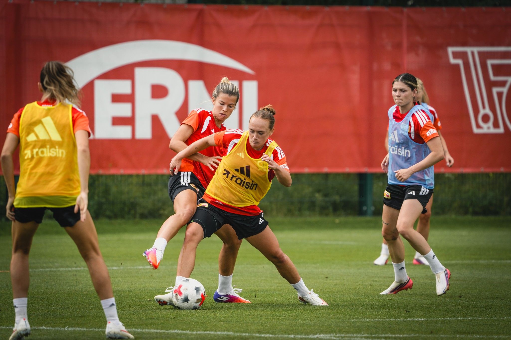 Vier Fußballspielerinnen trainieren auf einem Rasenplatz. Eine Spielerin dribbelt den Ball, während eine andere sie anrempelt.