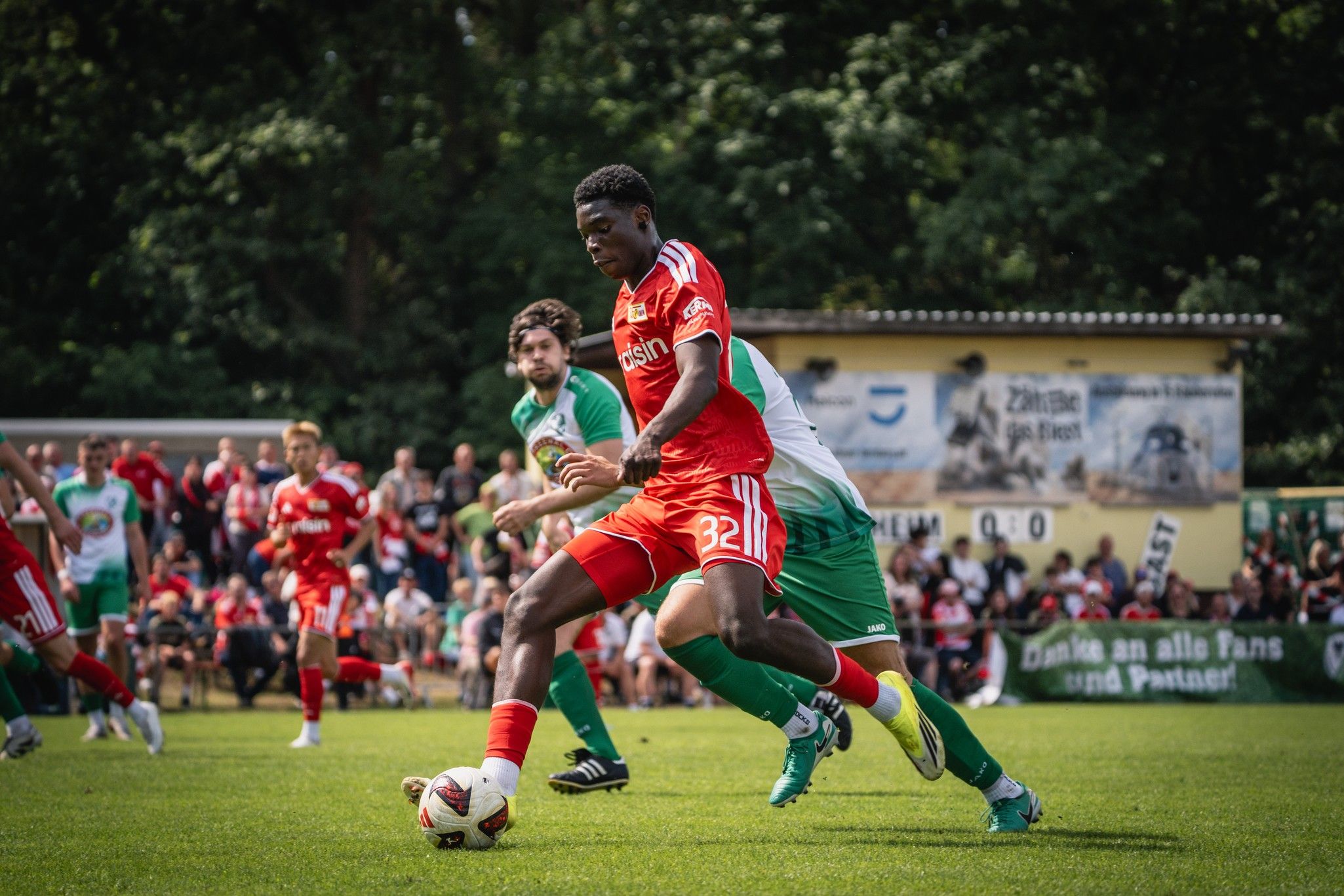 Ein Fußballspieler in roter Uniform dribbelt den Ball, während ein gegnerischer Spieler in grüner Uniform ihn verfolgt. Zuschauer im Hintergrund.