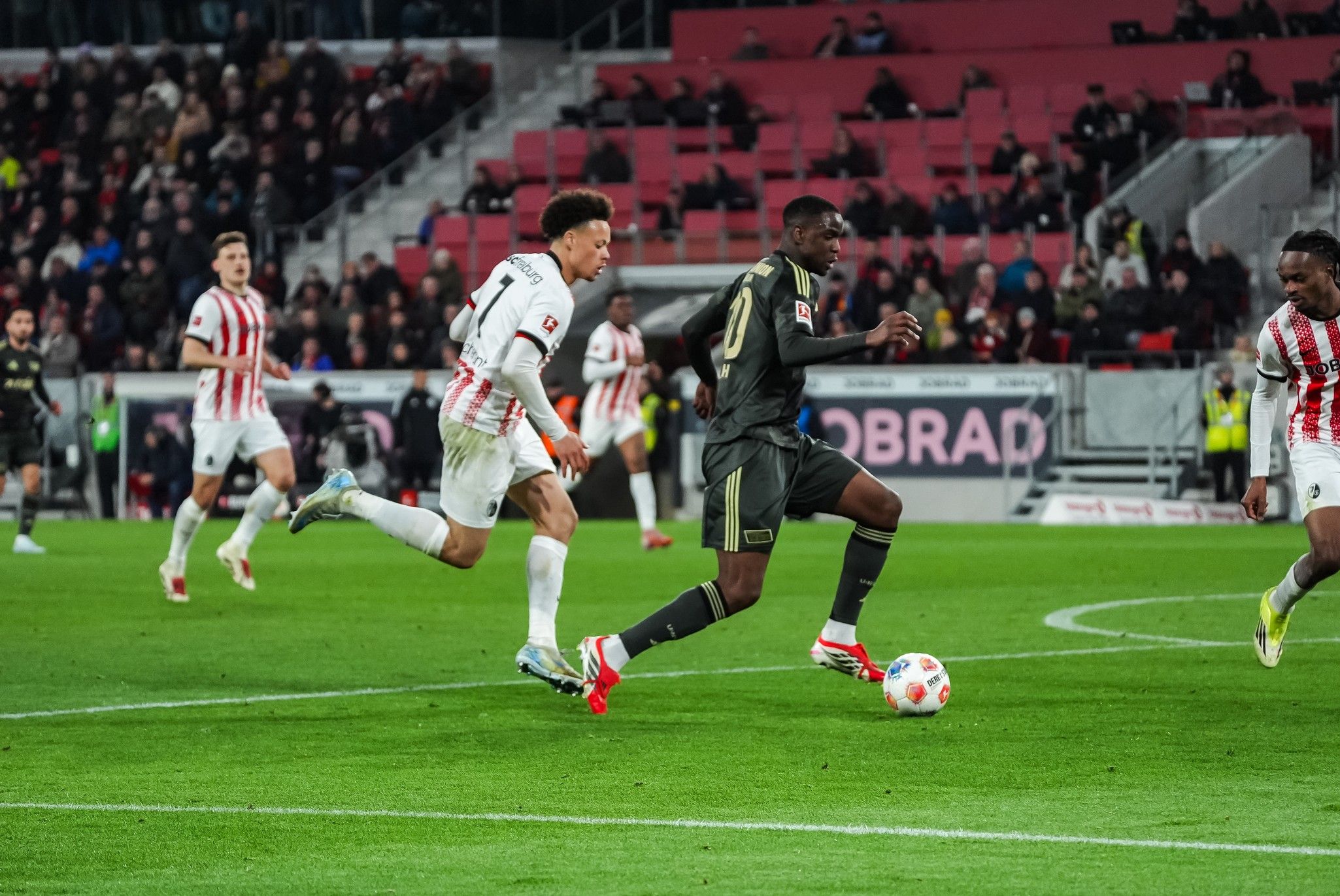 Ein Fußballspieler in dunkler Ausrüstung sprintet mit dem Ball, während ein Gegner ihn verfolgt. Zuschauer im Hintergrund.