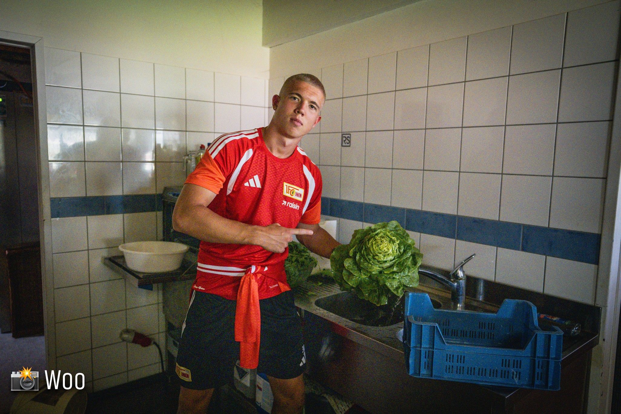 Ein junger Mann in einem roten Sportshirt steht in einer Küche und zeigt auf einen frischen Kopf Salat.