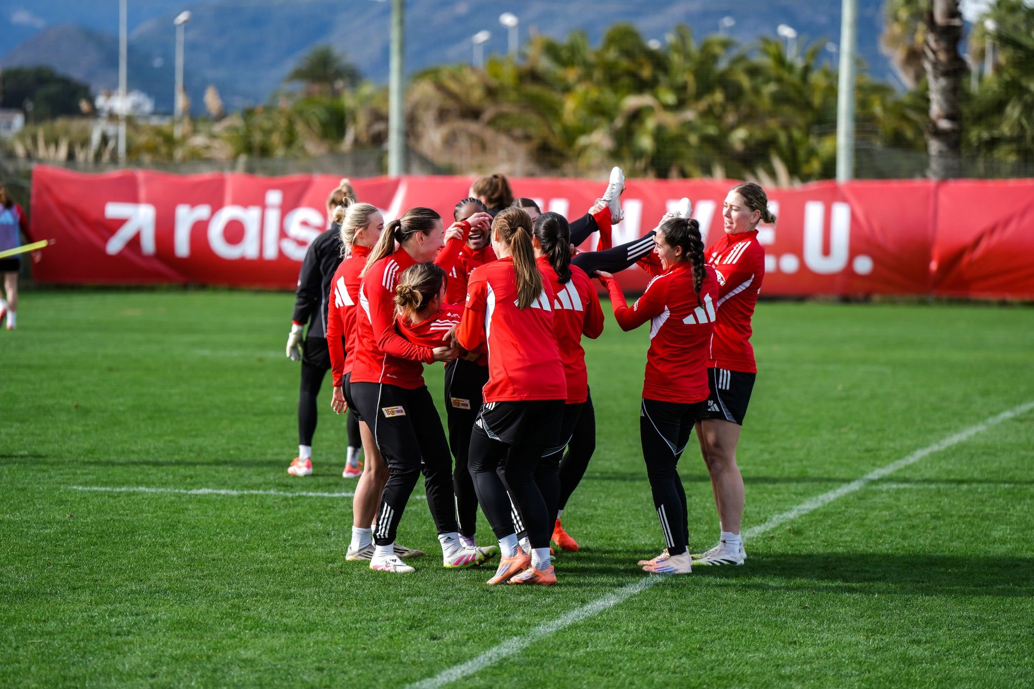 Mannschaftstraining auf dem Rasen, Spielerinnen in roten Trikots, die jubeln und sich gegenseitig unterstützen.