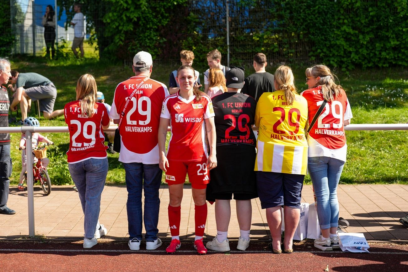 Gruppe von Fußballfans in roten und gelben Trikots mit der Nummer 29, stehen an einer Sportanlage.