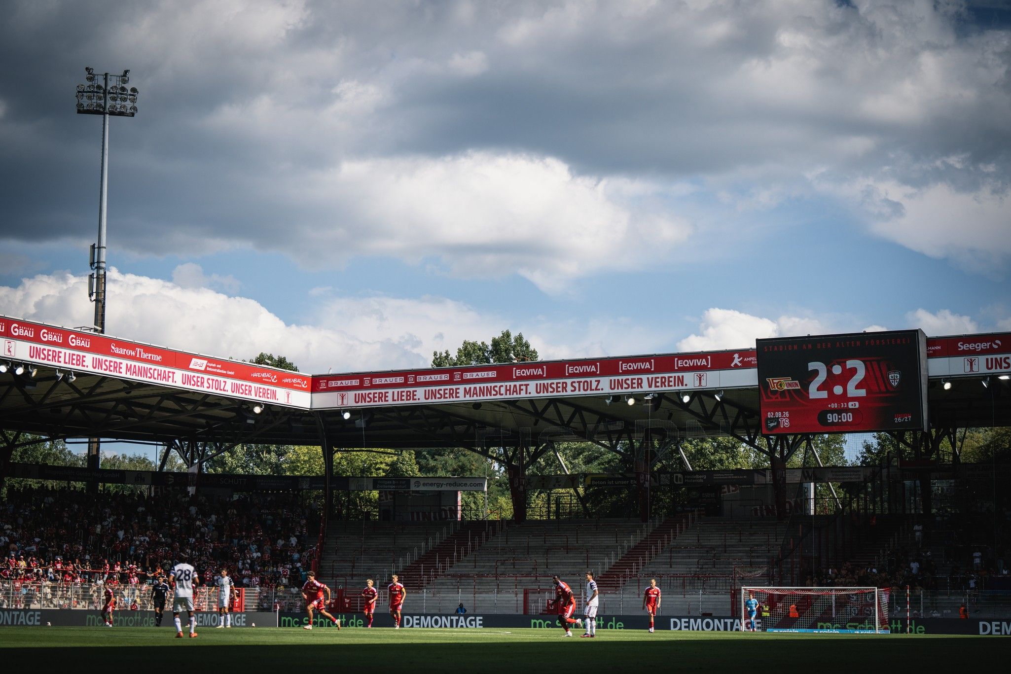Spielszene im Stadion mit einem Fußballspiel, zwei Mannschaften auf dem Platz und dem Stand von 2:2 auf der Anzeigetafel.