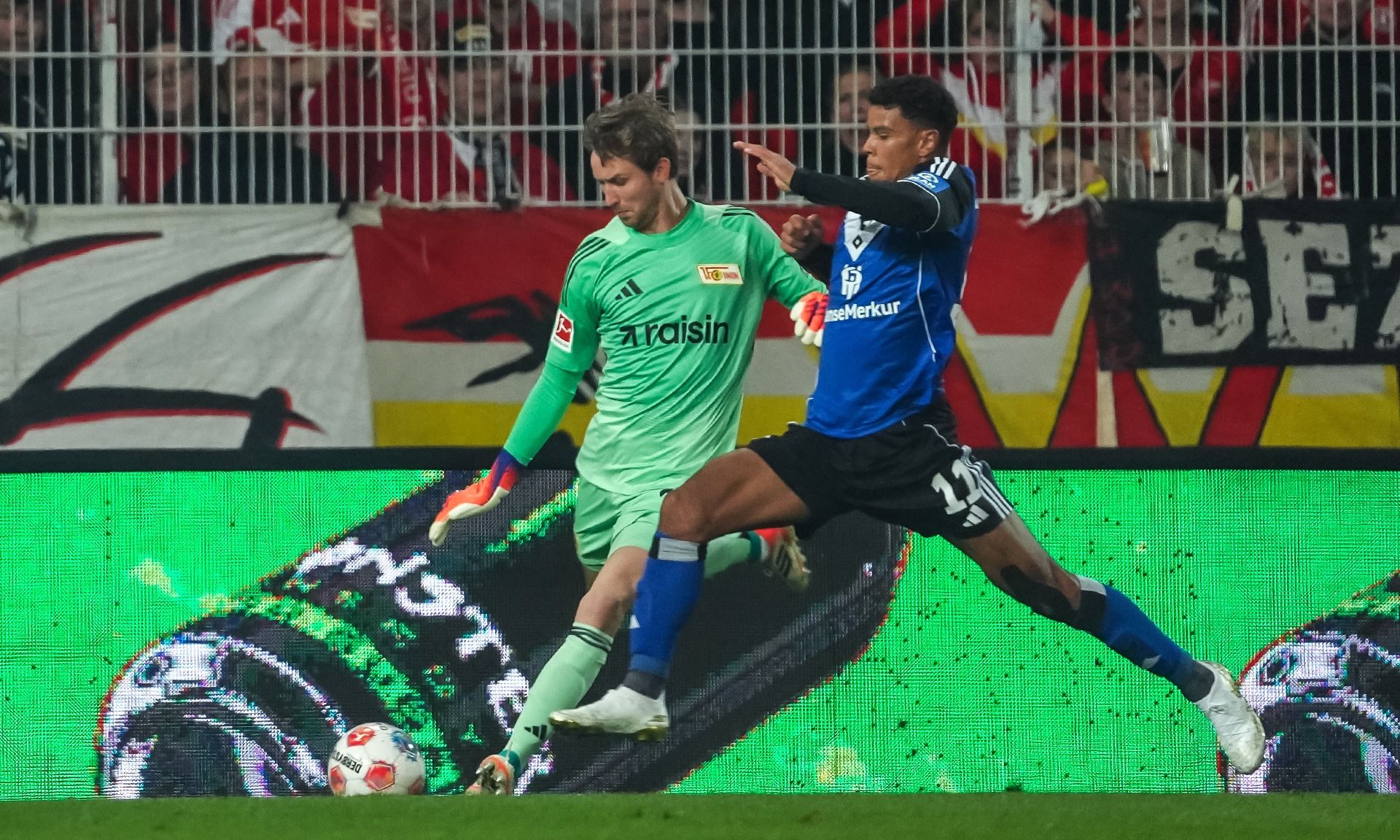 Torwart in grün weicht einem angreifenden Spieler in blau aus, während im Hintergrund Fanbanner zu sehen sind.