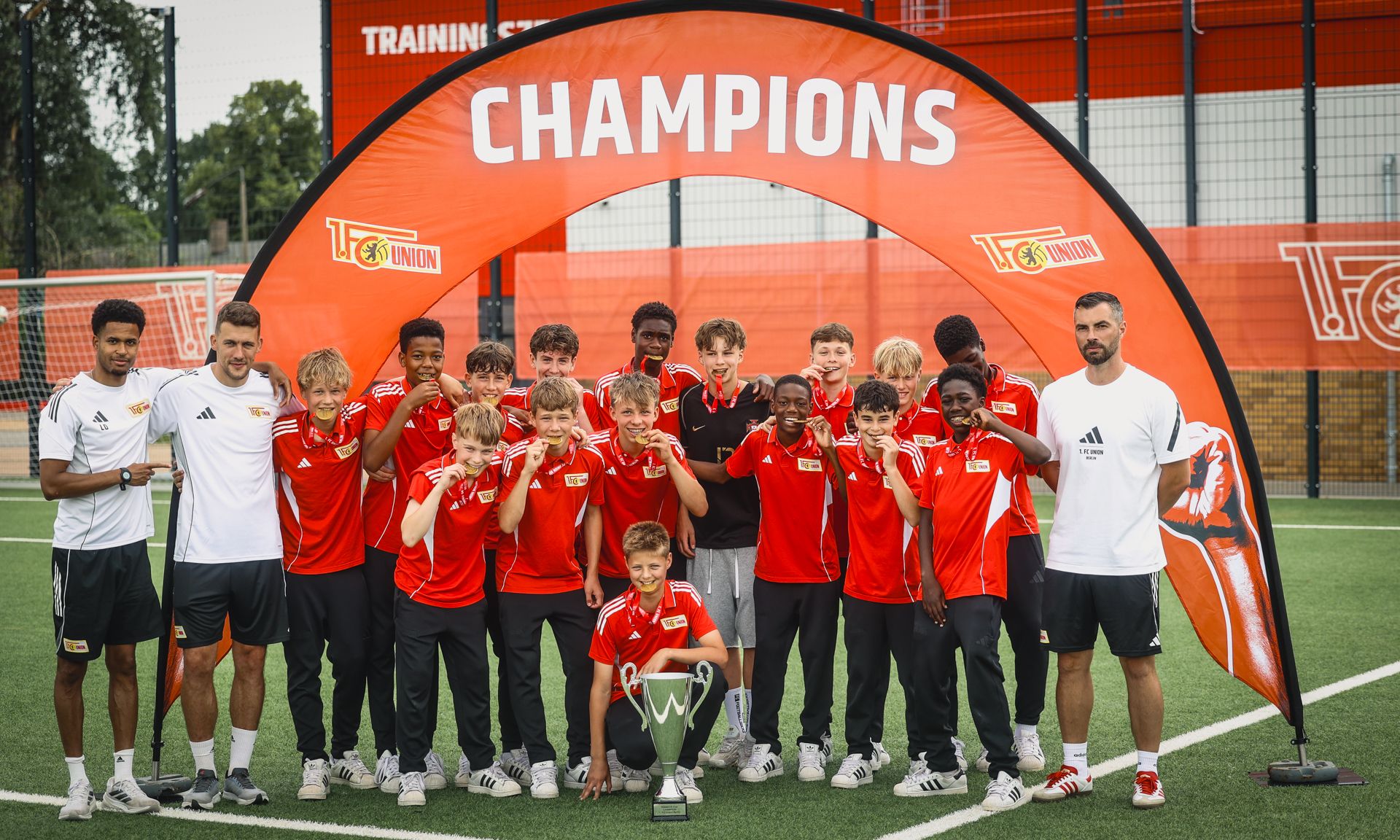 Gruppe von jungen Fußballspielern in roten Trikots posiert stolz mit einem Pokal unter einem "Champions"-Banner.
