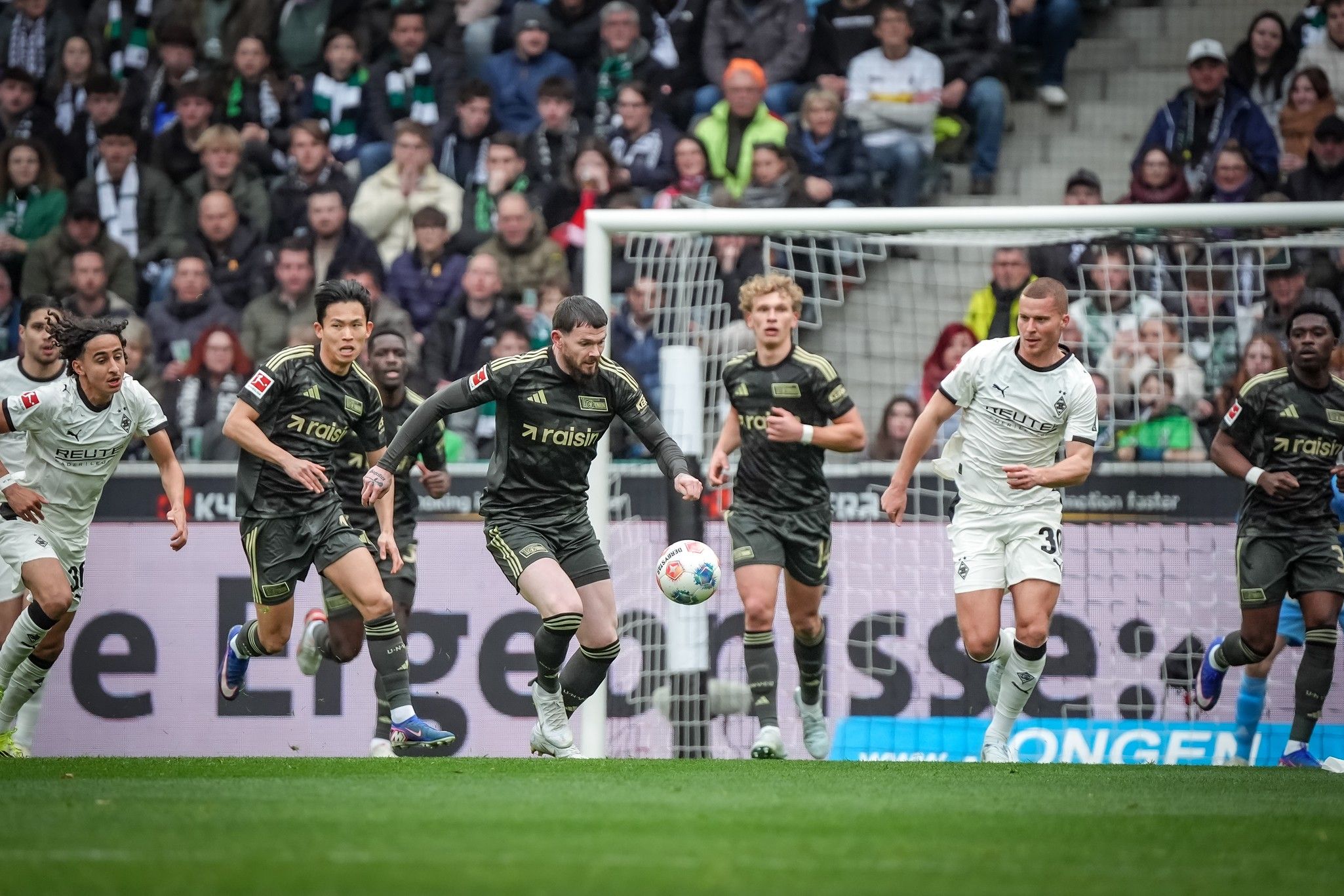 Spielszene eines Fußballspiels, in der mehrere Spieler um den Ball kämpfen, während Zuschauer im Hintergrund zuschauen.