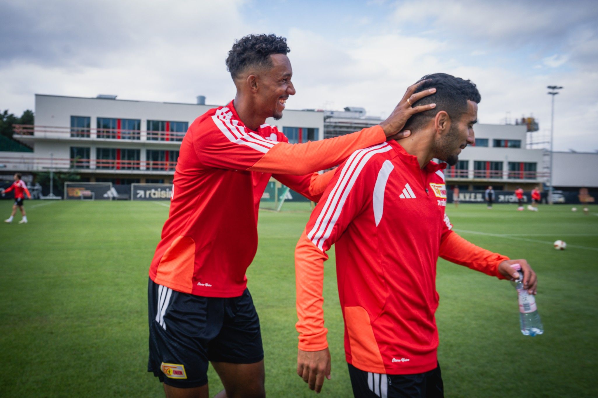 Zwei Fußballspieler im Training, einer berührt den Kopf des anderen lächelnd, beide tragen rote Trikots.