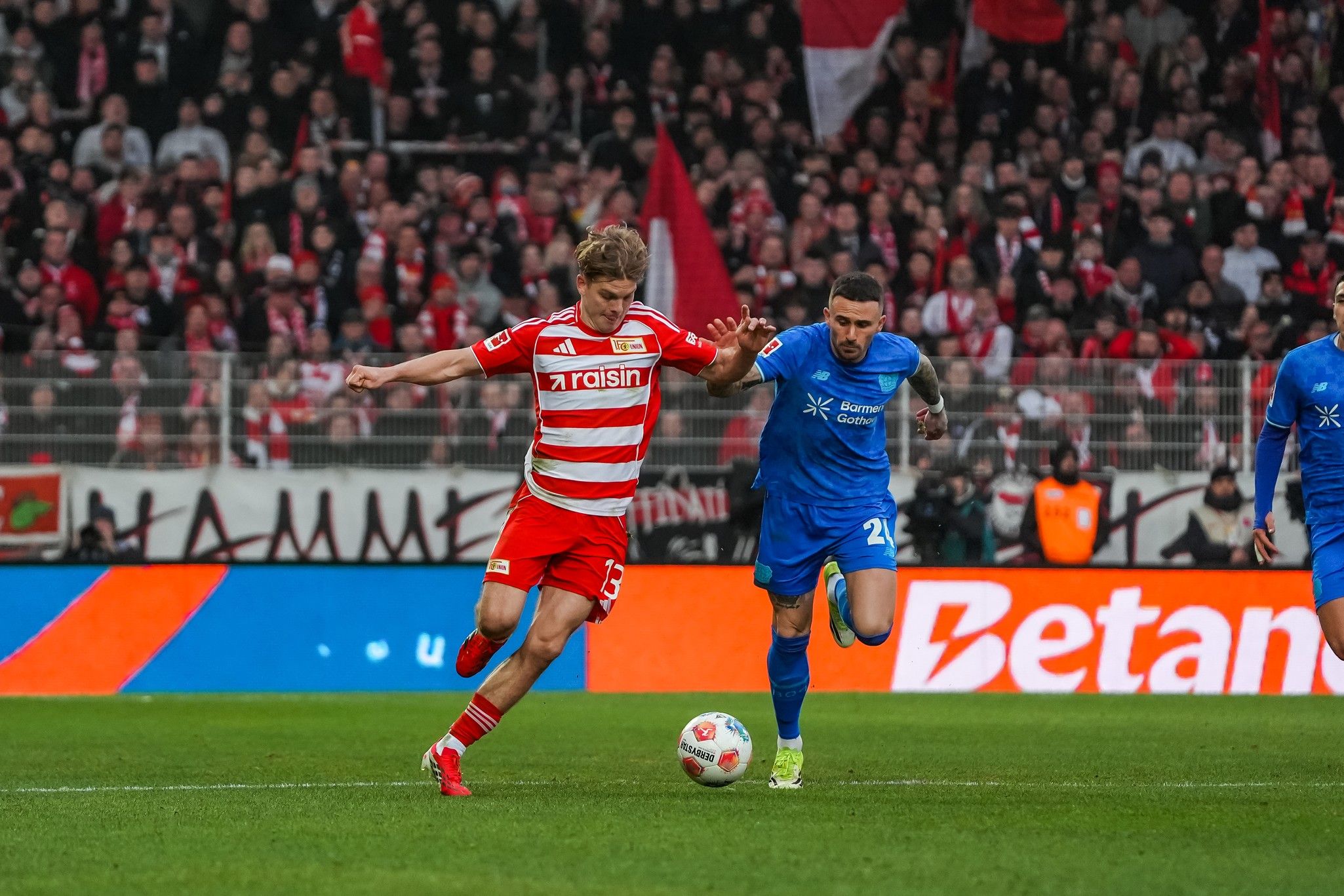 Ein Spieler in rot-weißer Uniform dribbelt den Ball, während ein Gegner in blauer Uniform ihn verfolgt. Im Hintergrund Fans im Stadion.