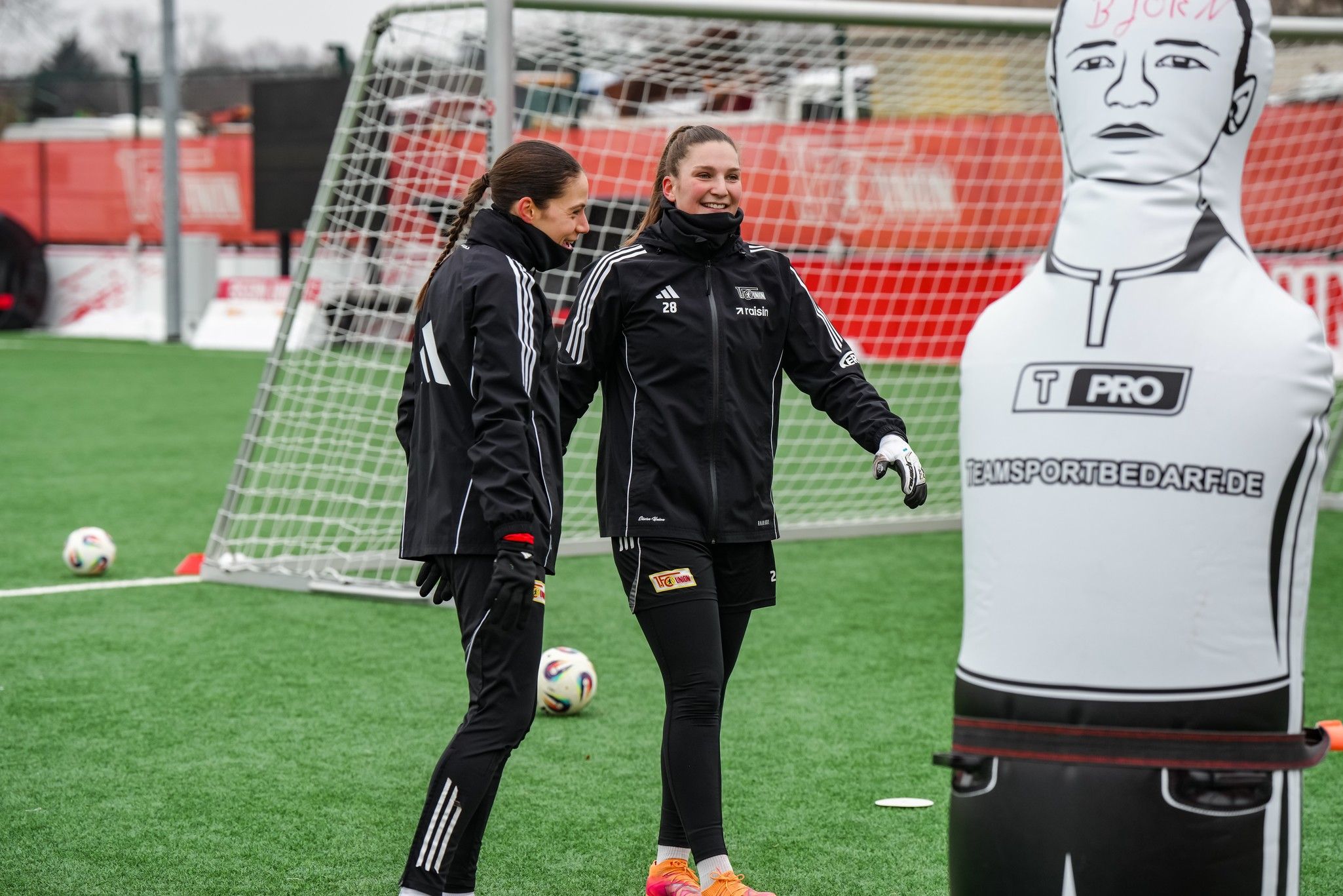 Zwei Fußballspielerinnen trainieren auf einem Platz neben einer Trainingspuppe vor einem Tor.