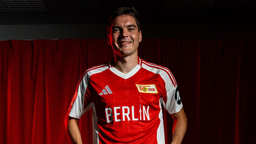 Ein Fußballspieler in einem roten Trikot mit der Aufschrift "Berlin" steht vor einem roten Hintergrund und lächelt.