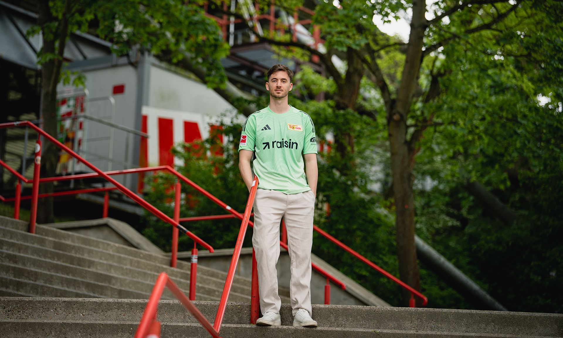 Mann in grünem Trikot und beigen Hosen steht auf einer Treppe mit rotem Geländer in einem parkähnlichen Umfeld.