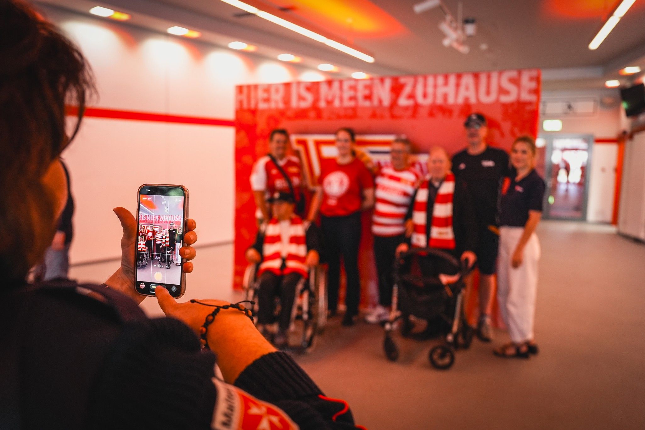 Eine Gruppe von Menschen posiert vor einem Banner mit der Aufschrift "Hier ist mein Zuhause", während jemand ein Foto mit dem Handy macht.