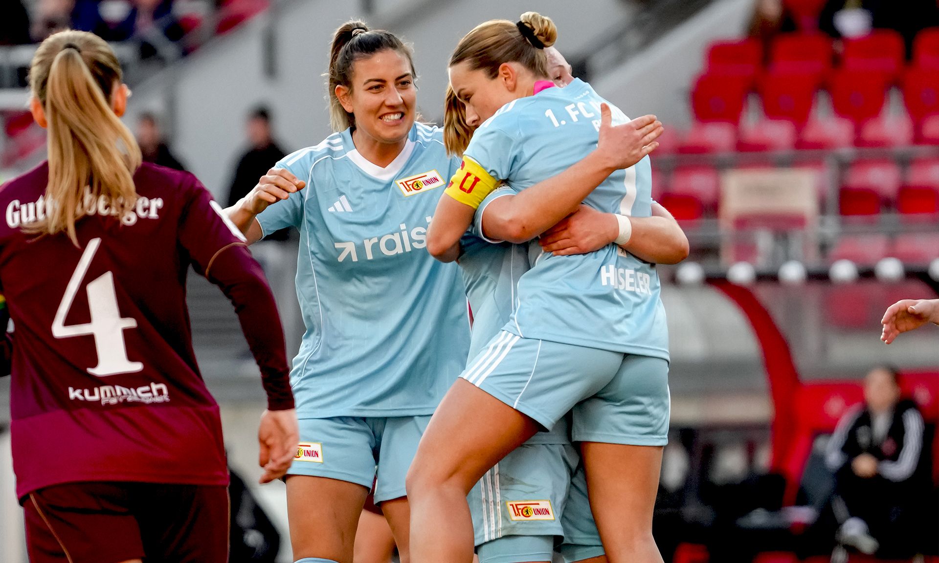 Drei Fußballspielerinnen in blauen Trikots umarmen sich jubelnd auf dem Spielfeld.