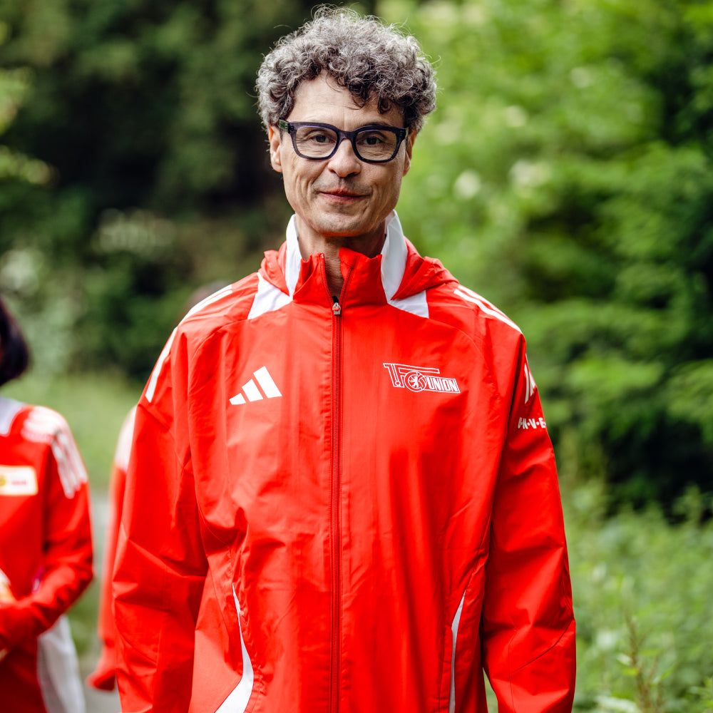 Ein Mann hat lockiges Haar und eine Brille. Er trägt eine rote Jacke mit einem Logo. Er steht in einer grünen Umgebung.