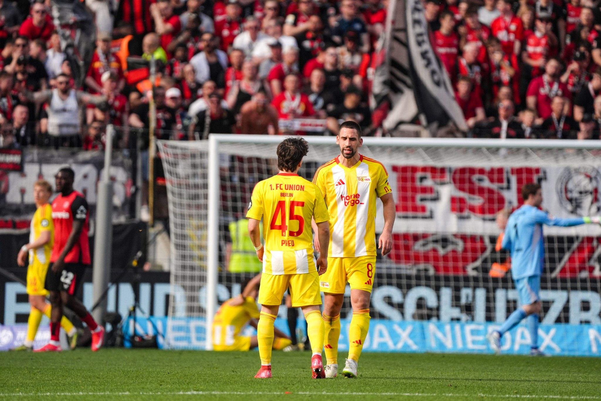 Zwei Fußballspieler des 1. FC Union Berlin in gelb-roten Trikots stehen auf dem Spielfeld, umgeben von jubelnden Fans.