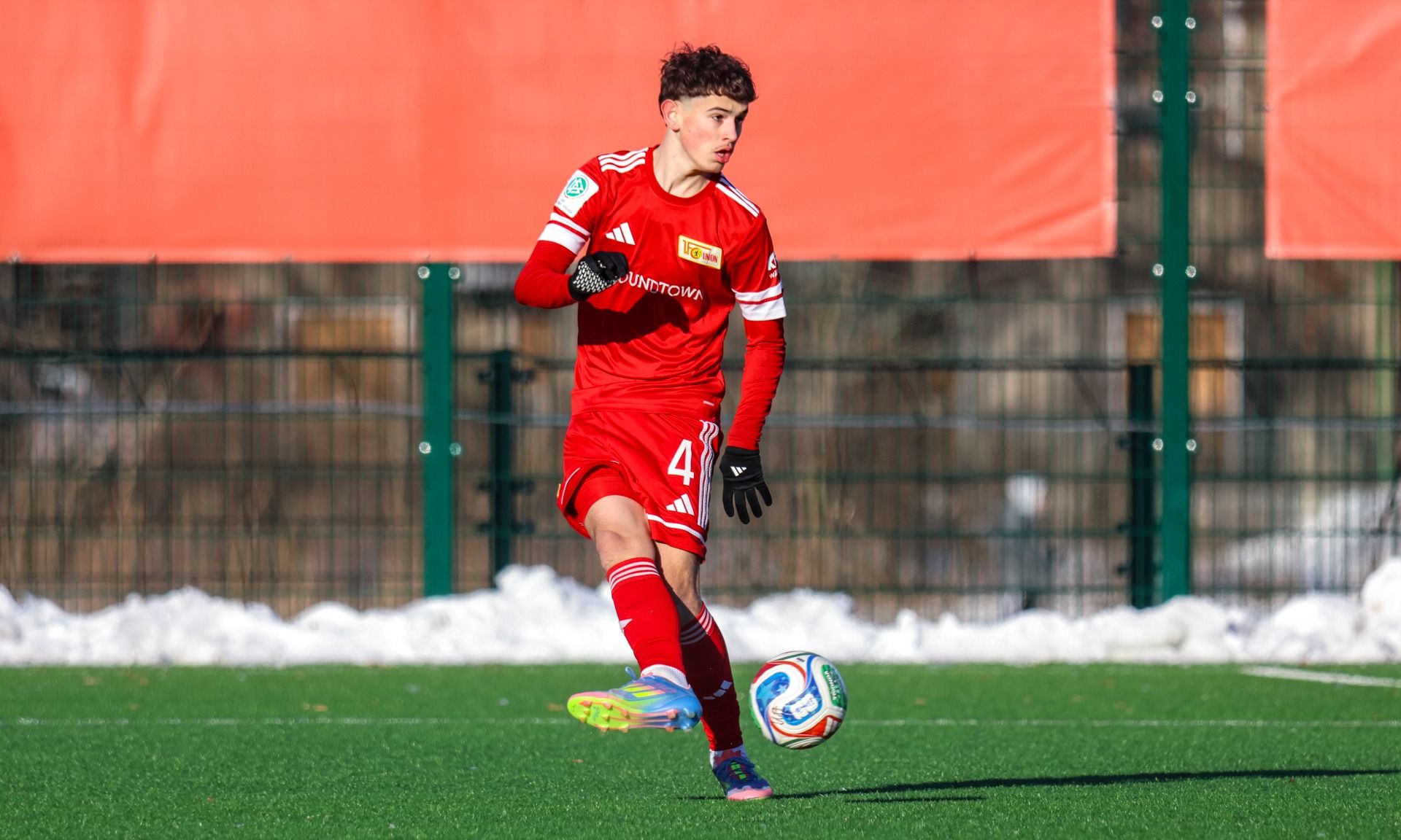 Ein junger Fußballspieler in roter Ausrüstung dribbelt den Ball auf einem Kunstrasenplatz.