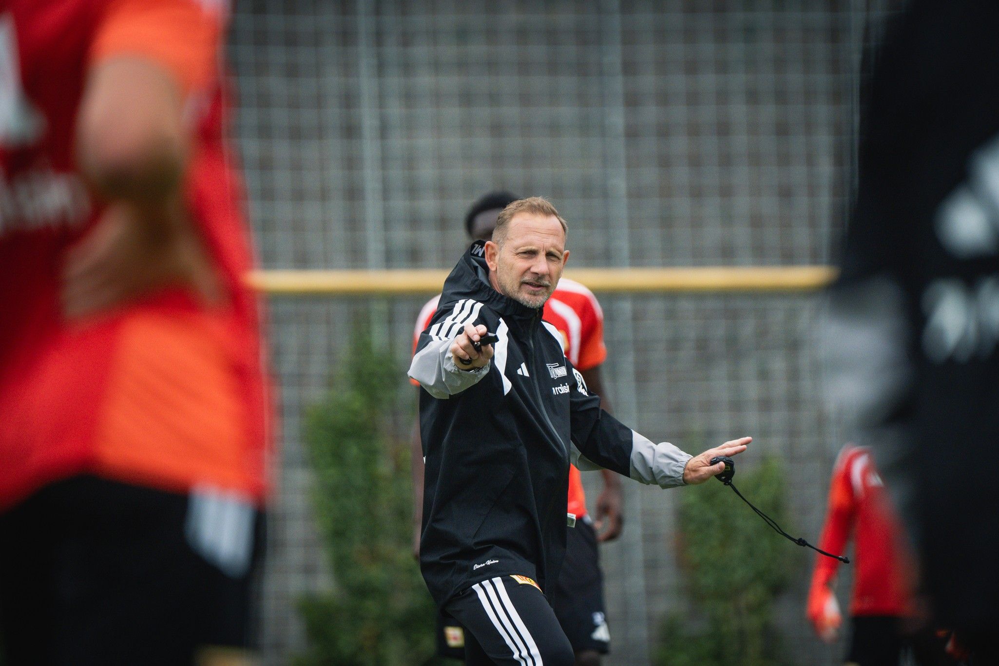 Ein Fußballtrainer beobachtet das Training und gibt Anweisungen an die Spieler in roten Trikots.