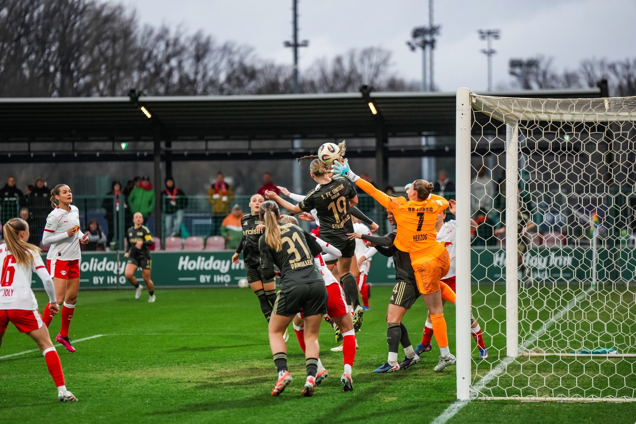Torwart fängt den Ball während eines Fußballspiels, mehrere Spielerinnen sind im Hintergrund aktiv.