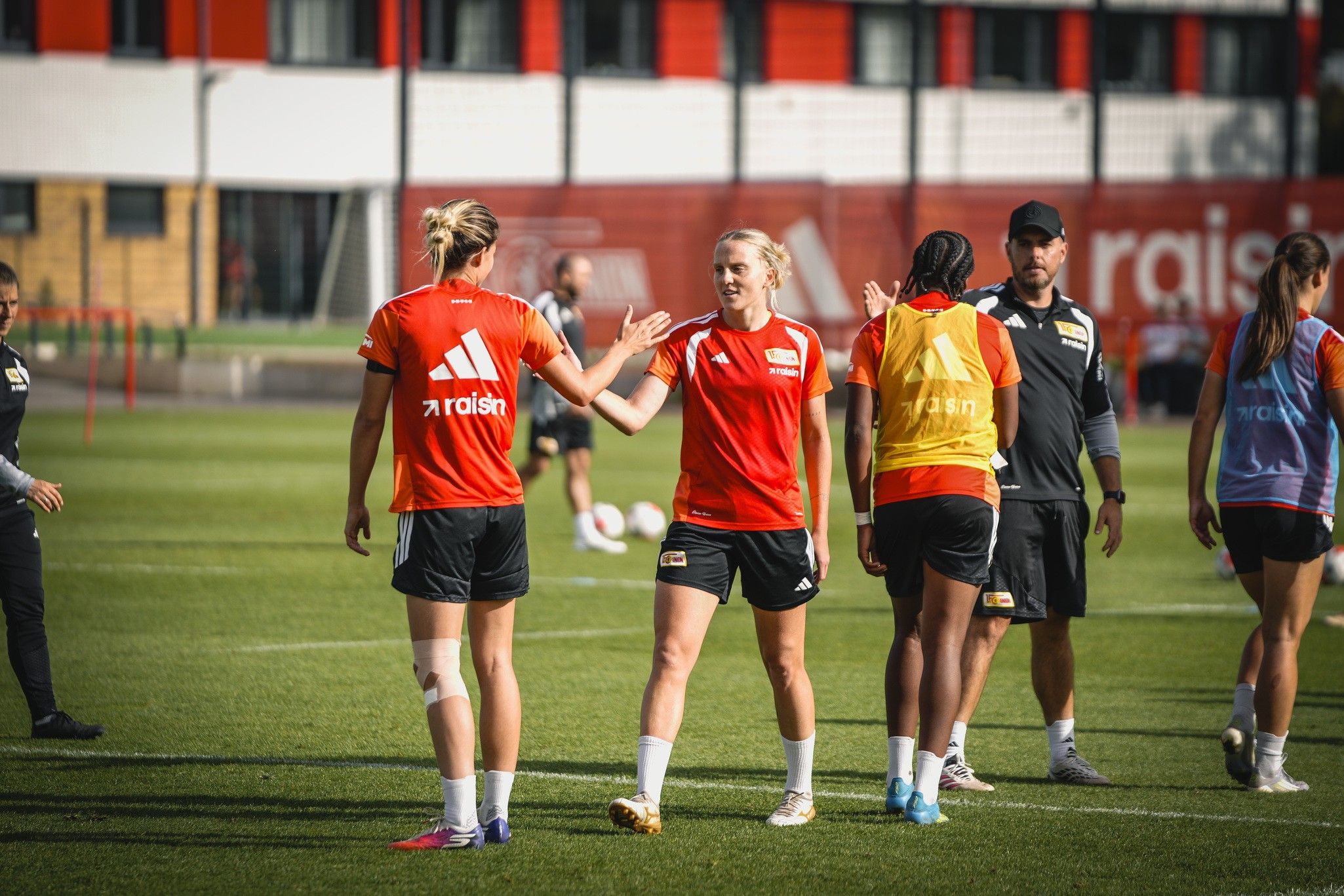 Zwei Frauen in roten Trainingsshirts begrüßen sich auf einem Fußballplatz, während ein Trainer im Hintergrund zuschaut.