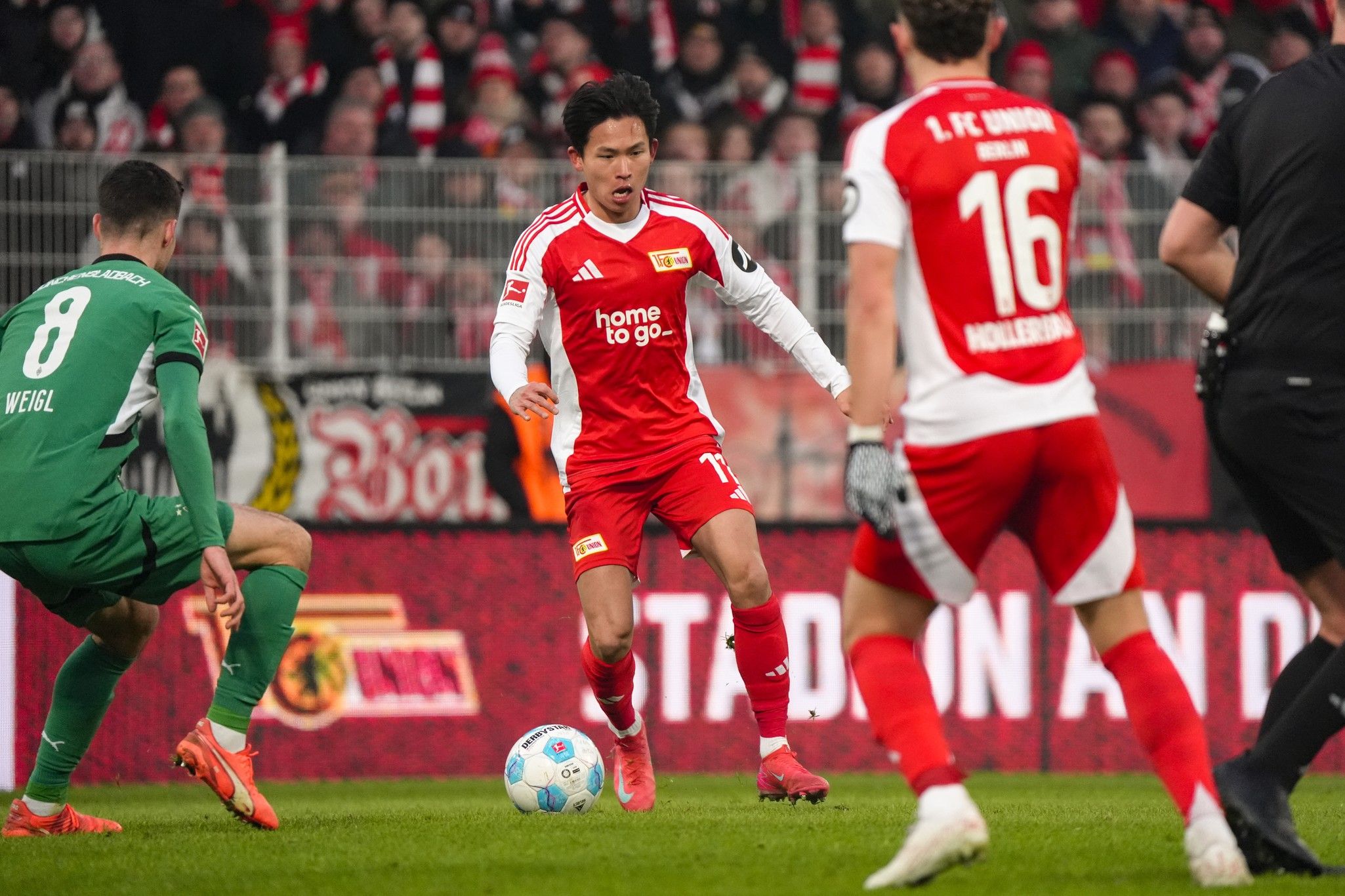 Ein Fußballspieler in rotem Trikot dribbelt den Ball, während zwei gegnerische Spieler und ein Torwart in der Nähe stehen.