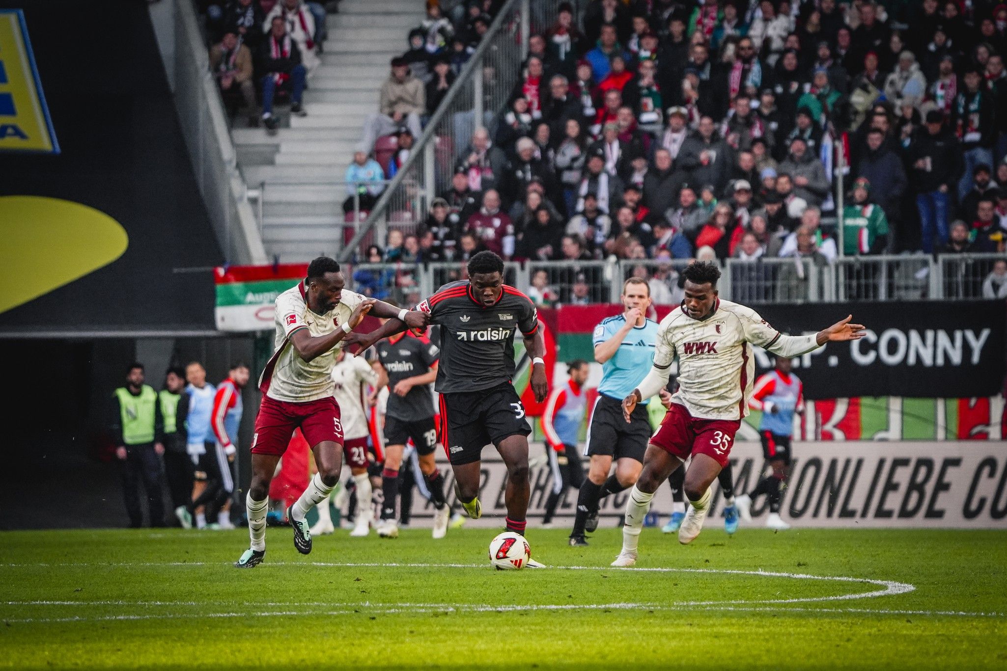 Ein Fußballspieler dribbelt den Ball, während zwei Verteidiger ihn verfolgen. Im Hintergrund ist ein Schiedsrichter und Fans zu sehen.