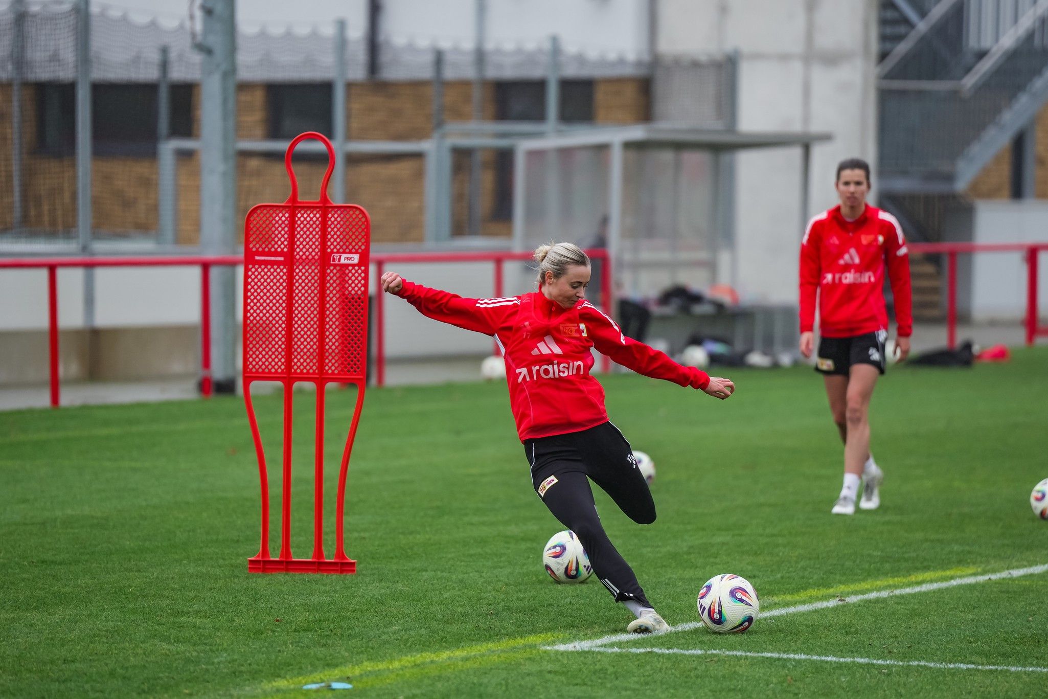Eine Spielerin in rotem Trikot trainiert auf dem Feld und schießt den Ball, während eine andere Spielerin im Hintergrund steht.