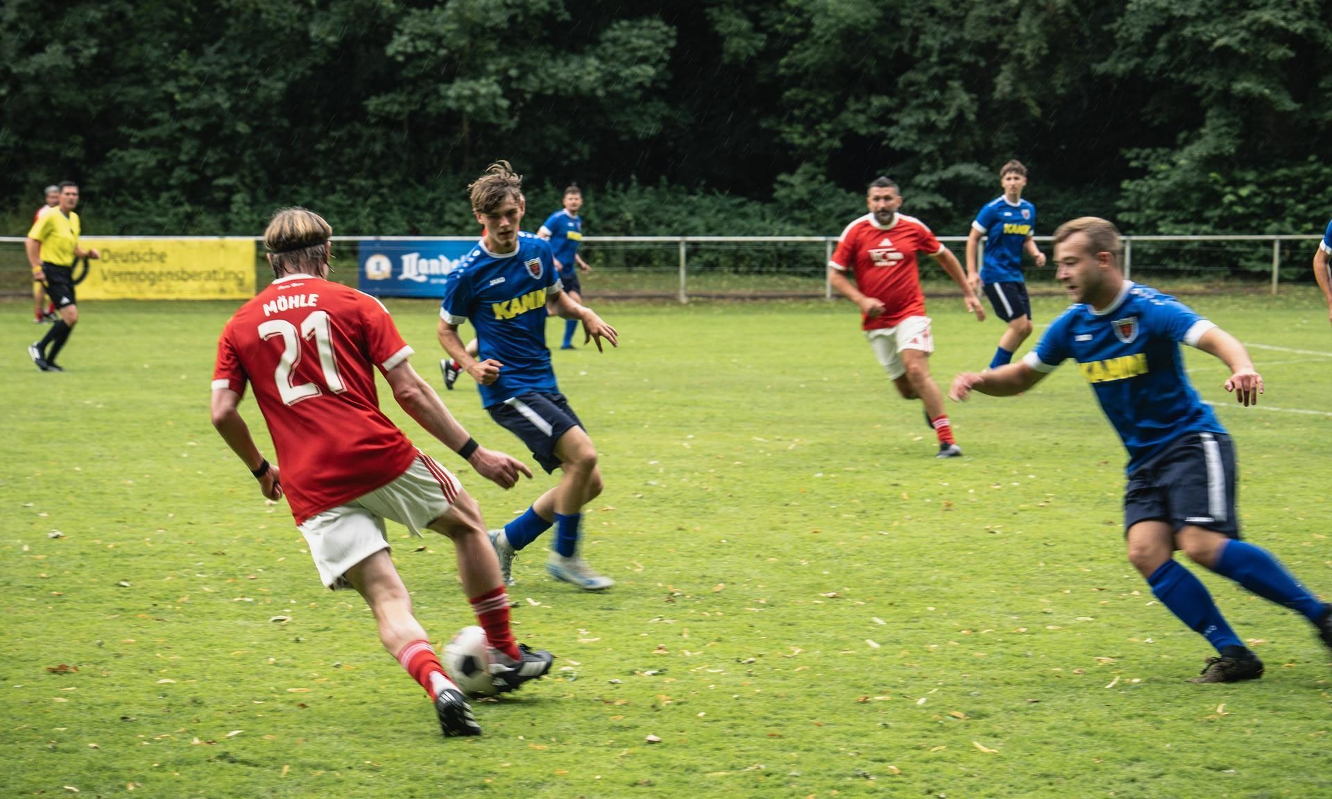Zwei Fußballspieler in roten und blauen Trikots stehen sich auf einem grünen Rasenfeld gegenüber, während weitere Spieler im Hintergrund agieren.