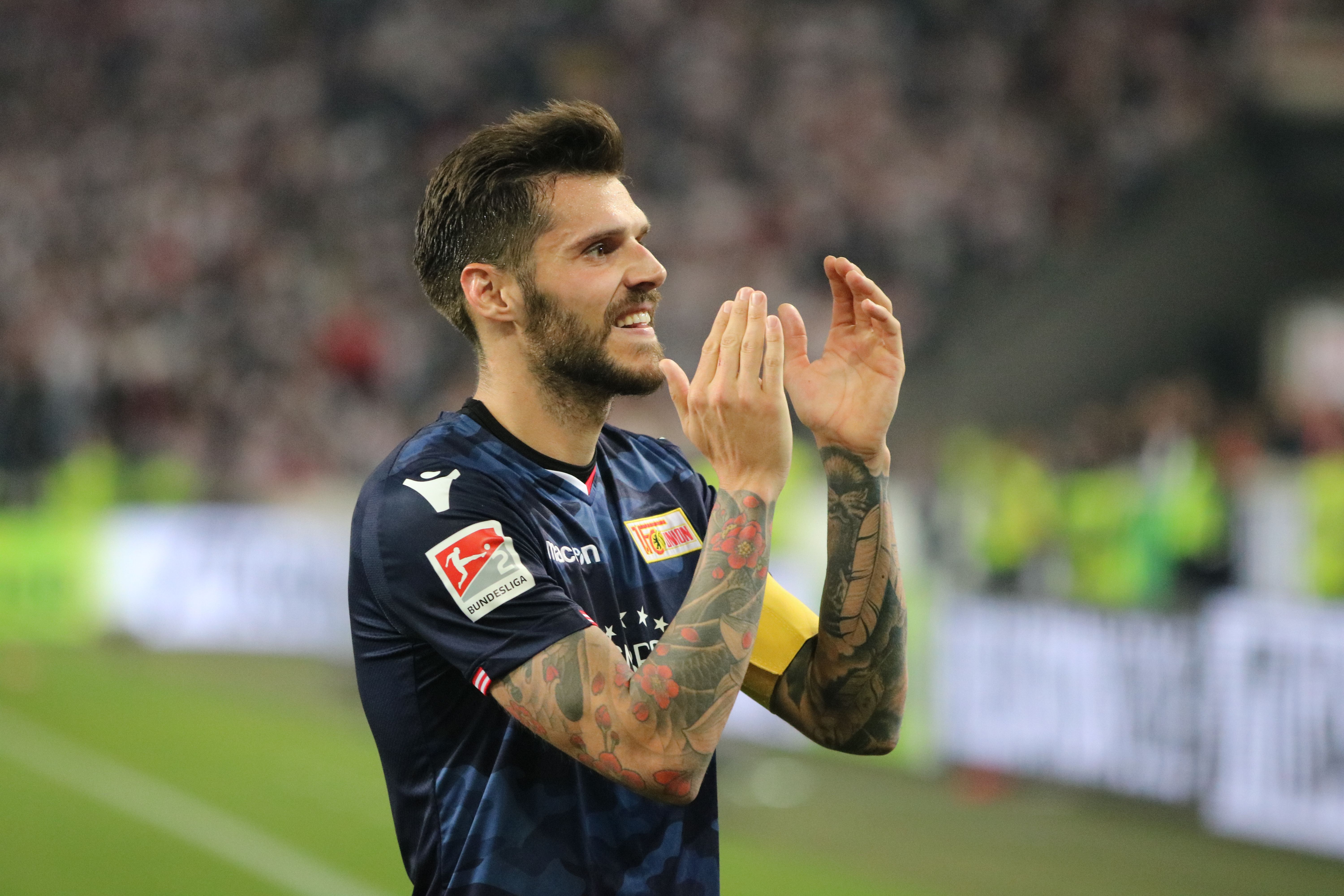 Ein Fußballspieler mit Tattoos applaudiert auf dem Spielfeld, trägt ein dunkles Trikot und hat eine Kapitänsbinde.