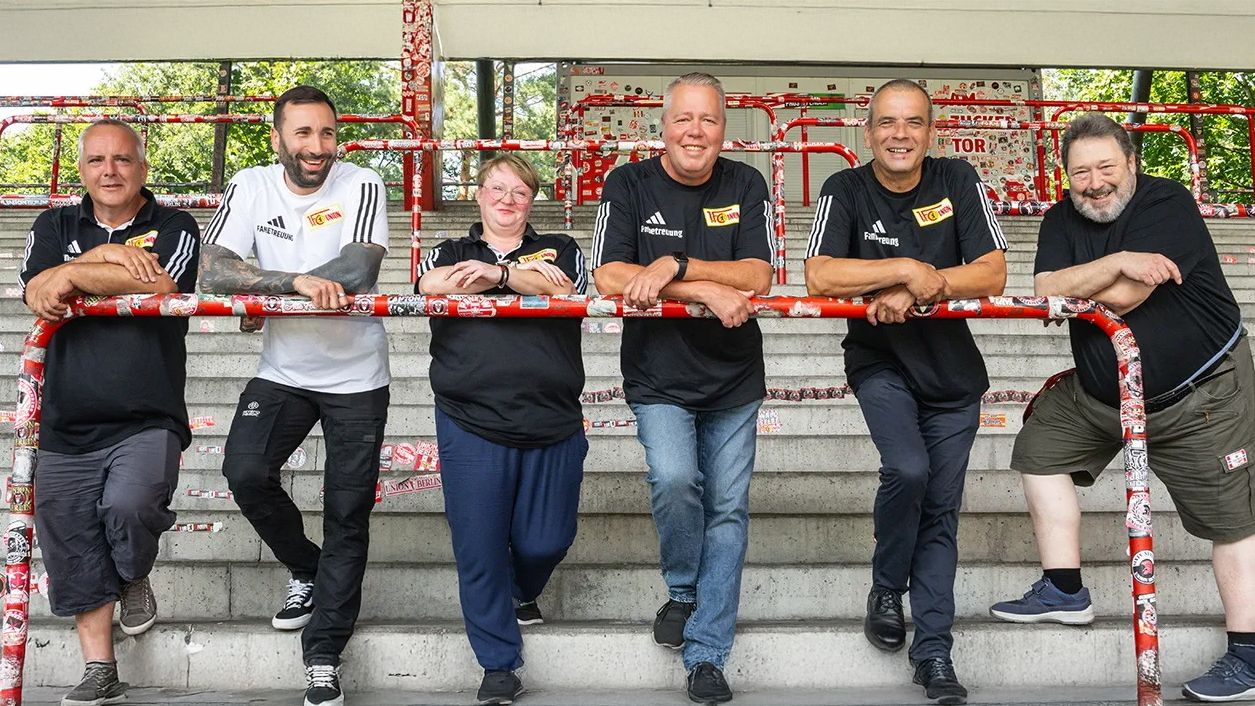 Sechs Personen stehen lächelnd an einem Geländer, gekleidet in schwarze T-Shirts, vor einer Sportanlage.