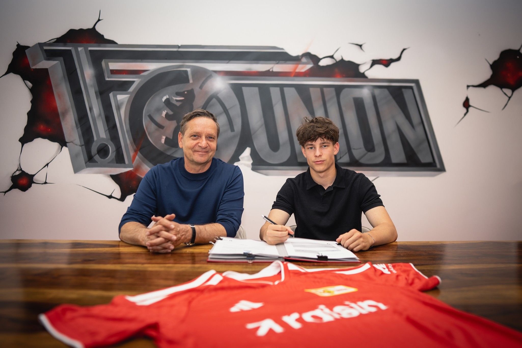 Ein Trainer und ein Spieler sitzen an einem Tisch mit Vertragsunterlagen und einem Fußballtrikot im Hintergrund.