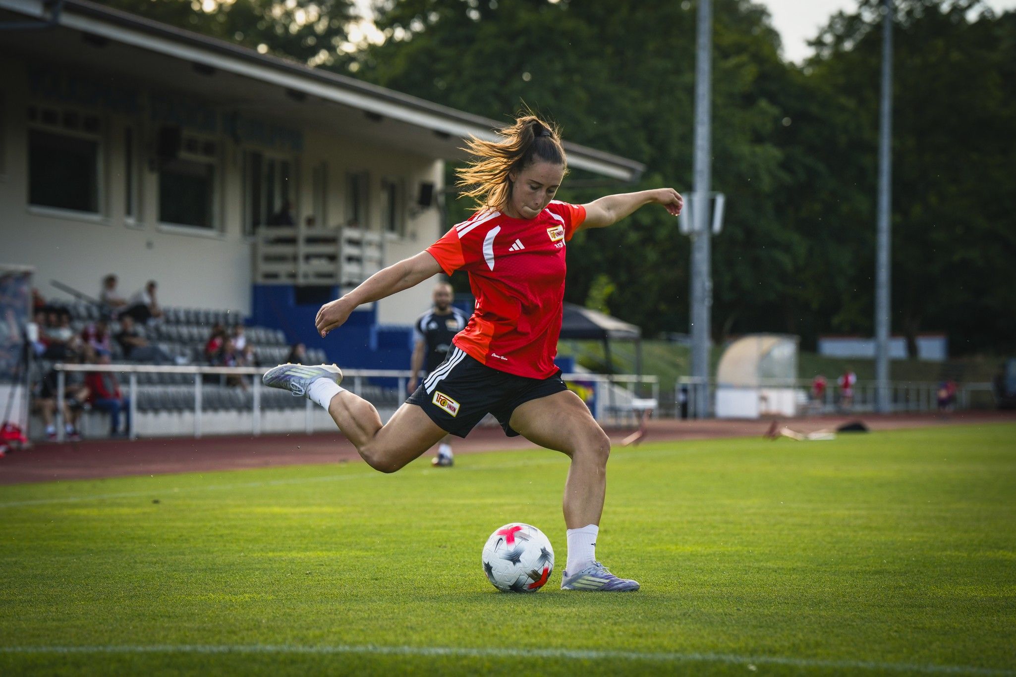 Eine Frau in rotem Trikot kickt einen Fußball auf einem Sportplatz. Im Hintergrund sind Zuschauer und Gebäude zu sehen.