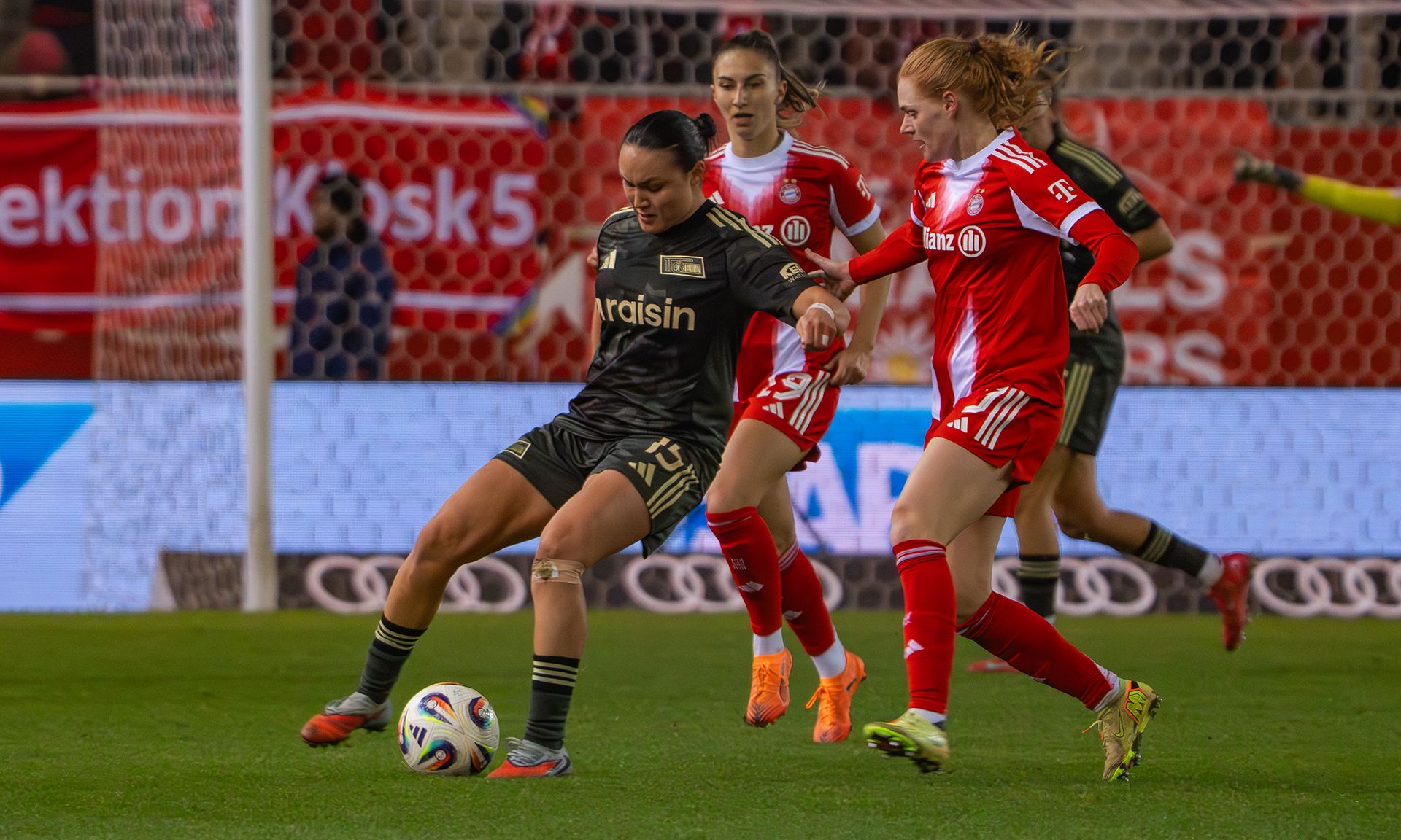 Eine Spielerin in schwarzem Trikot dribbelt den Ball, während zwei gegnerische Spielerinnen in roten Trikots sie drängen.