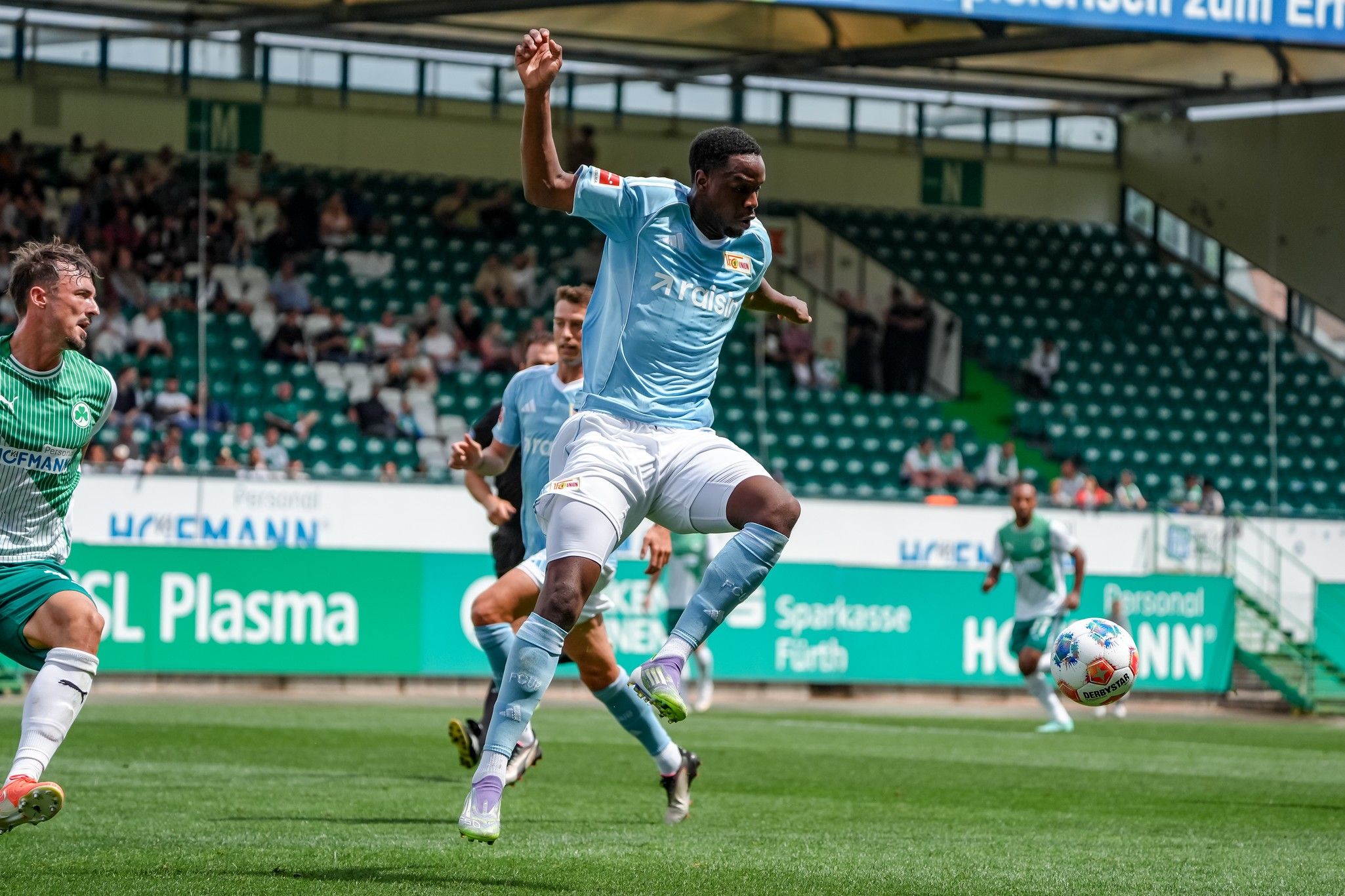 Spielszene im Fußball: Ein Spieler springt, während ein Gegner in grün-weißer Ausrüstung ihn anvisiert. Hintergrund mit Zuschauern im Stadion.