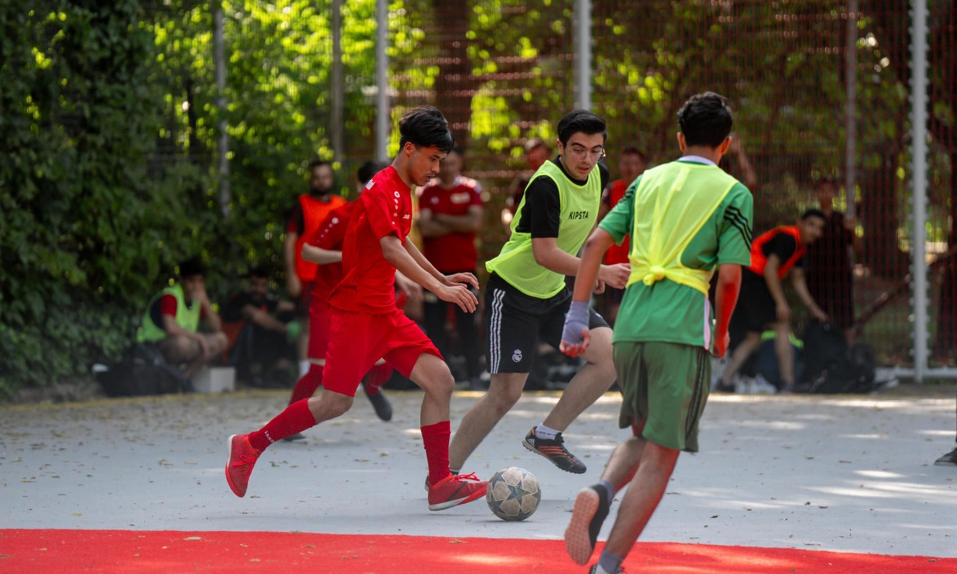 Drei junge Männer spielen Fußball auf einem Platz, der von Bäumen umgeben ist, während Zuschauer im Hintergrund zuschauen.