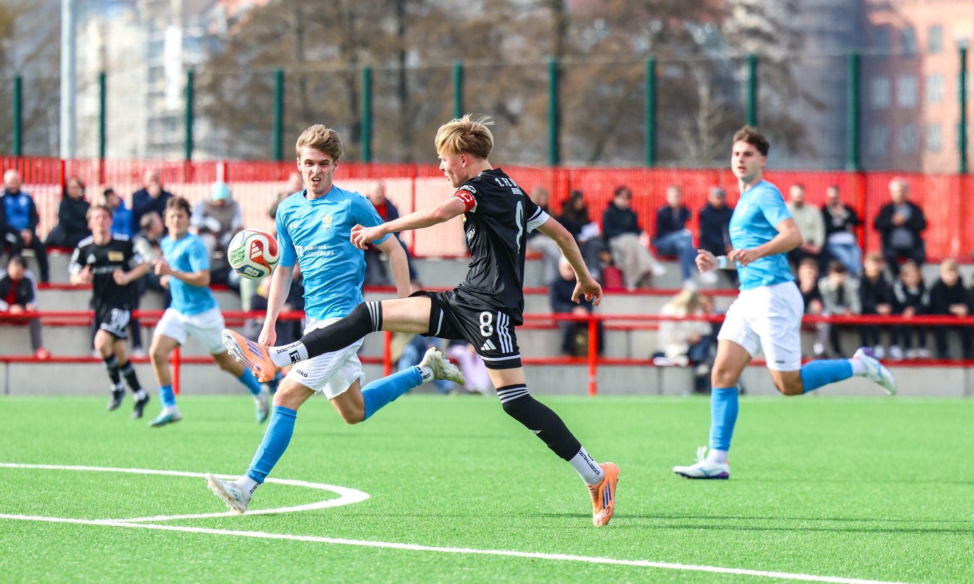 Zwei Fußballspieler im Wettkampf um den Ball auf einem Kunstrasenplatz, im Hintergrund Zuschauer.