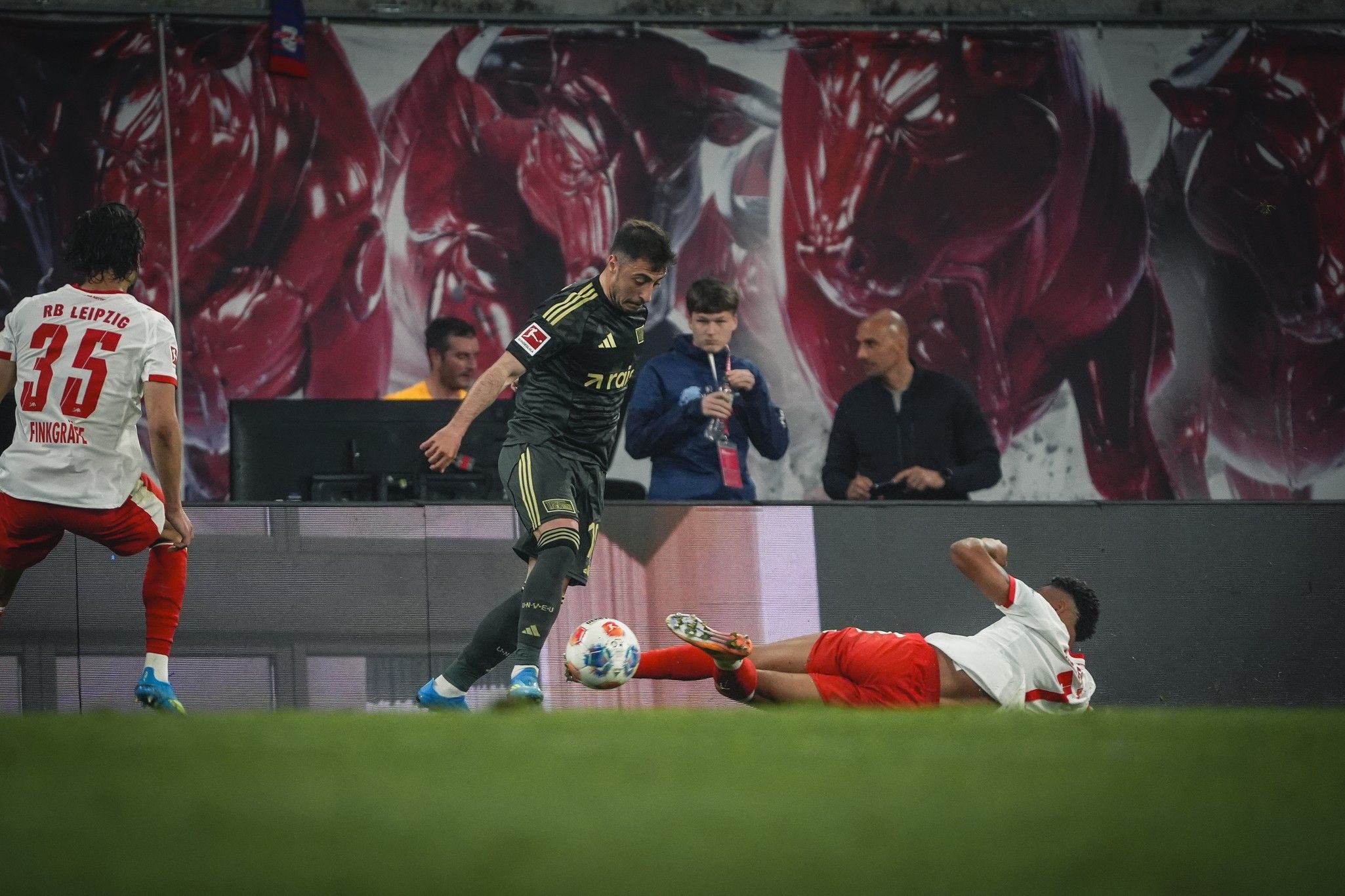 Ein Fußballspieler umgeht einen anderen, der am Boden liegt, während Zuschauer im Hintergrund zusehen.