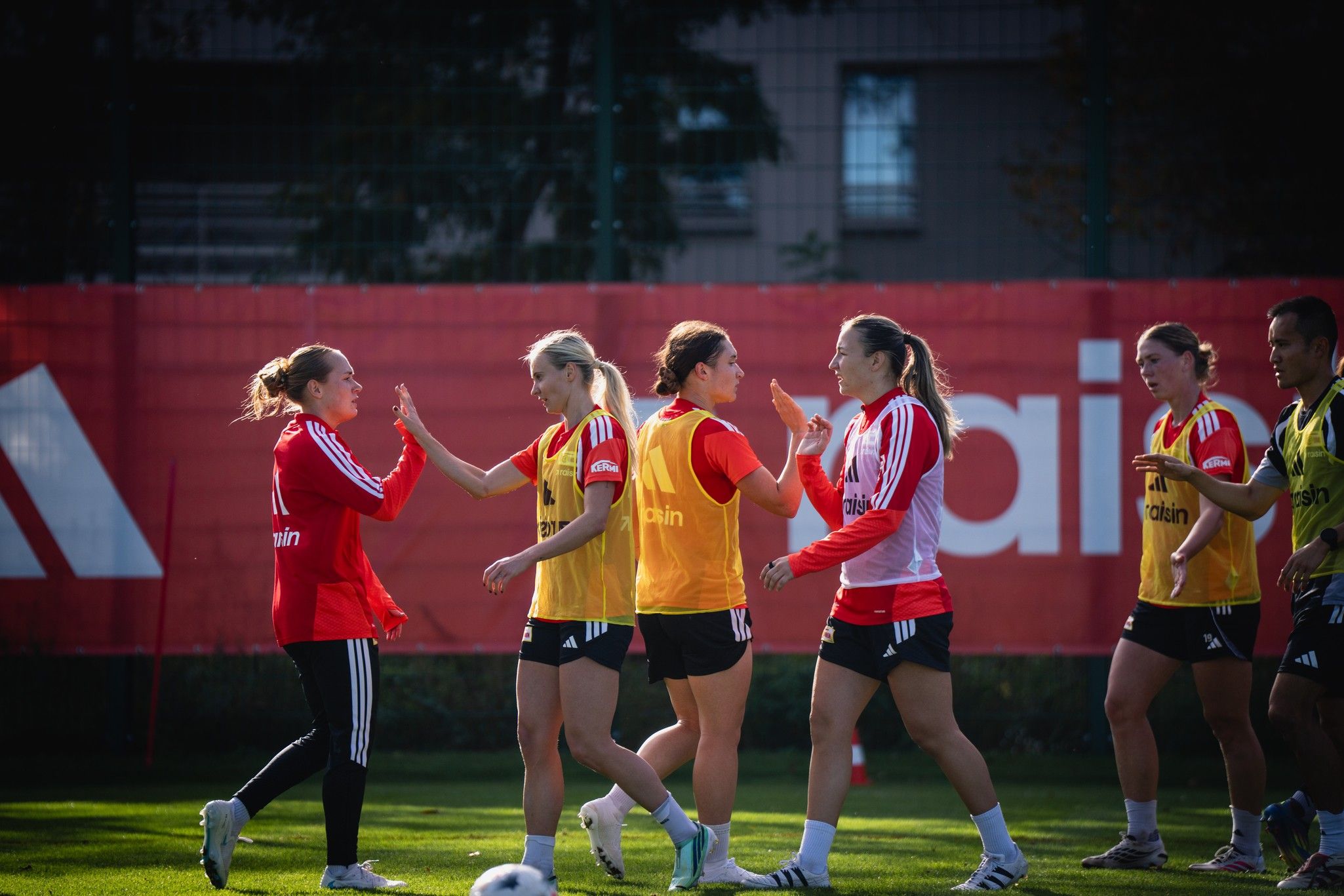 Frauenfußballmannschaft bei einem Training, Spielerinnen in roten und orangefarbenen Trikots, die sich begrüßen.
