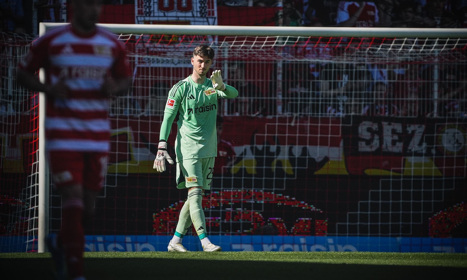 Ein Torwart in grünem Trikot steht im Tor und hebt die Hand zur Geste, während ein Spieler im Vordergrund vorbeiläuft.