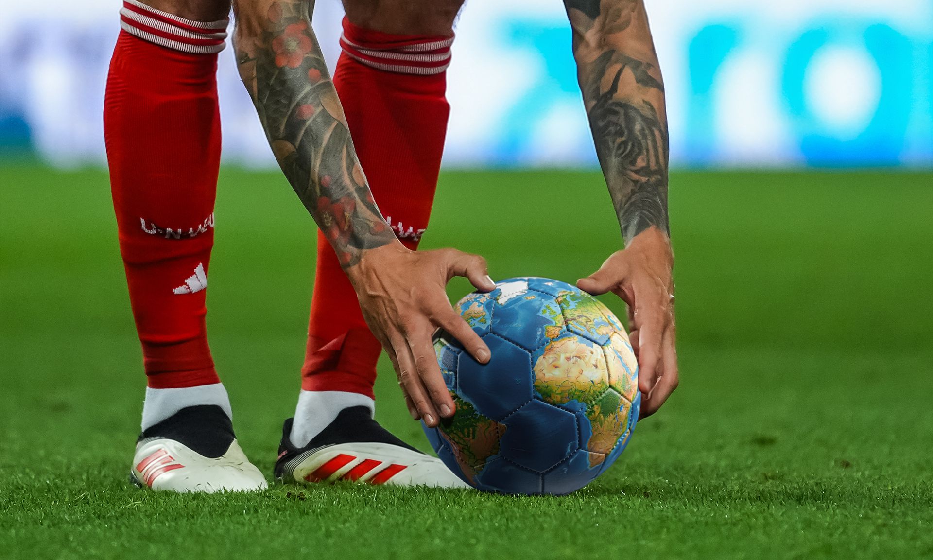 Ein Fußballspieler hält einen Fußball mit einer Weltkarte in den Händen, während er sich auf dem Spielfeld Positioniert.